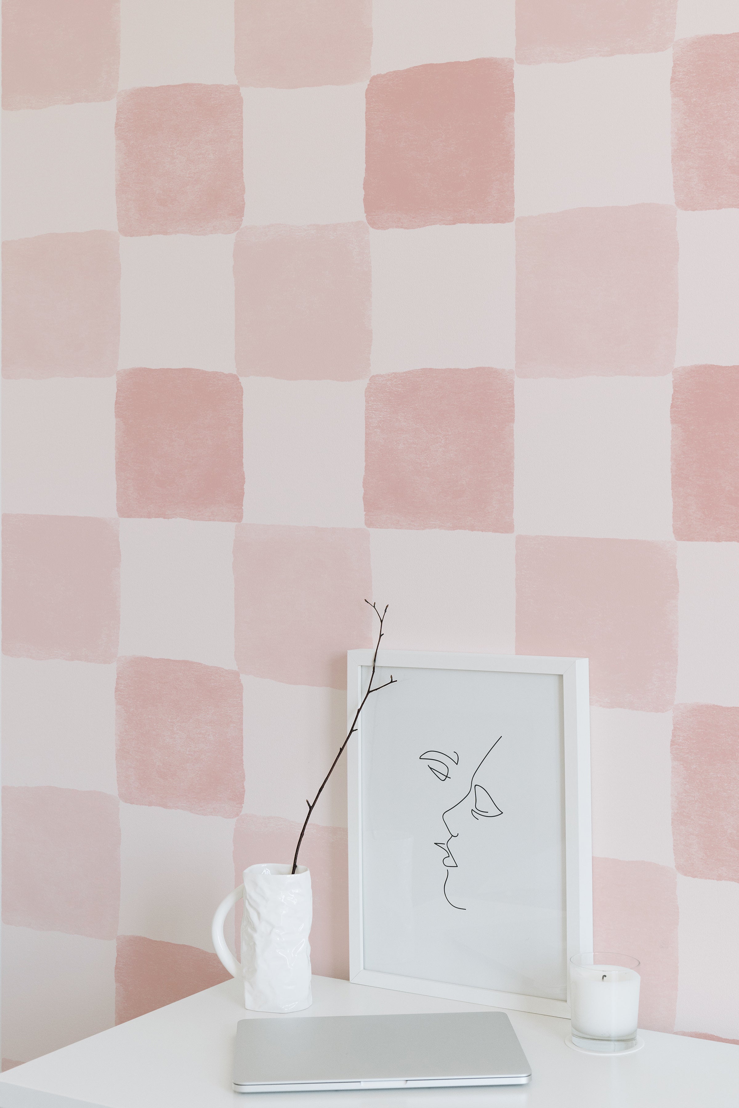 A close-up of the Clémence Wallpaper in Dusty Pink, showcasing a checkered pattern of large, soft dusty pink and white squares in a watercolor finish, with a minimalistic framed line drawing of a face on a white background, accompanied by a twig in a sculpted white vase and a lit candle.