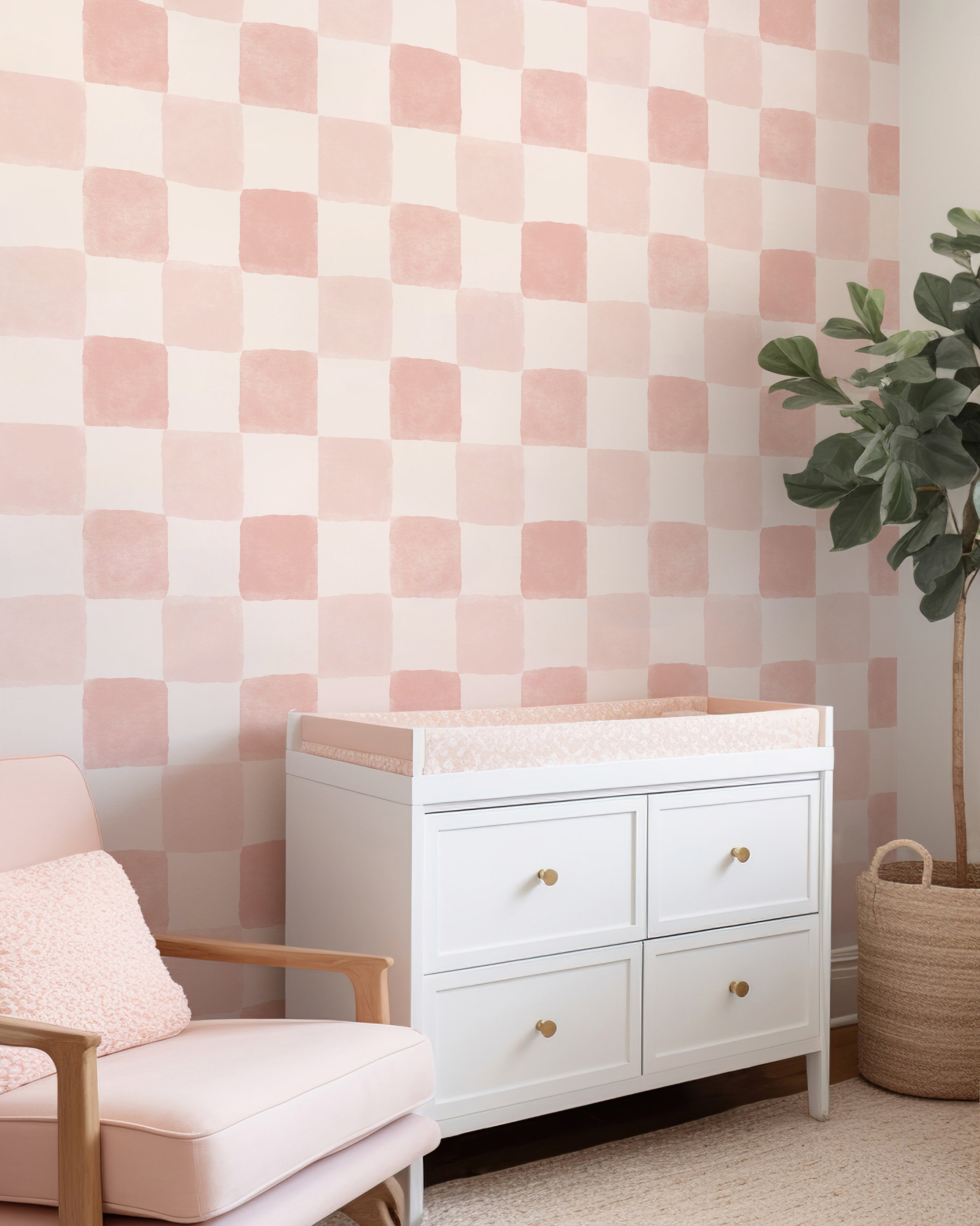 A stylish room setting featuring the Clémence Wallpaper in Dusty Pink with large checkered squares. The room is furnished with a white changing table, a pale pink armchair, and a potted green plant, offering a calm and elegant nursery space