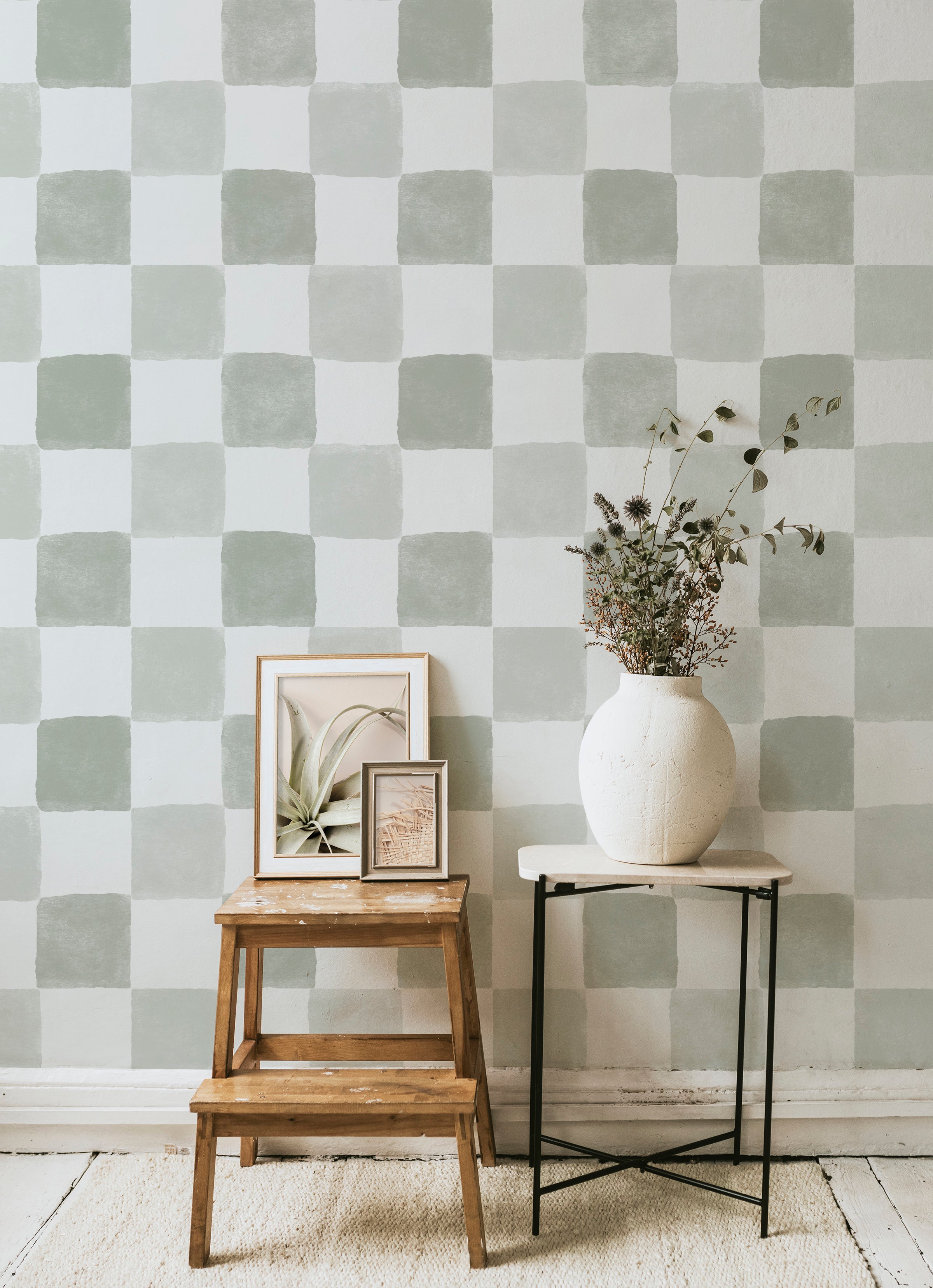 A stylish corner of a room showcasing the Clémence Wallpaper - Olive, a seamless checkered pattern. A rustic wooden step stool holds framed artwork and a large vase with dried flowers, enhancing the wallpaper's soft olive hues and handmade aesthetic.