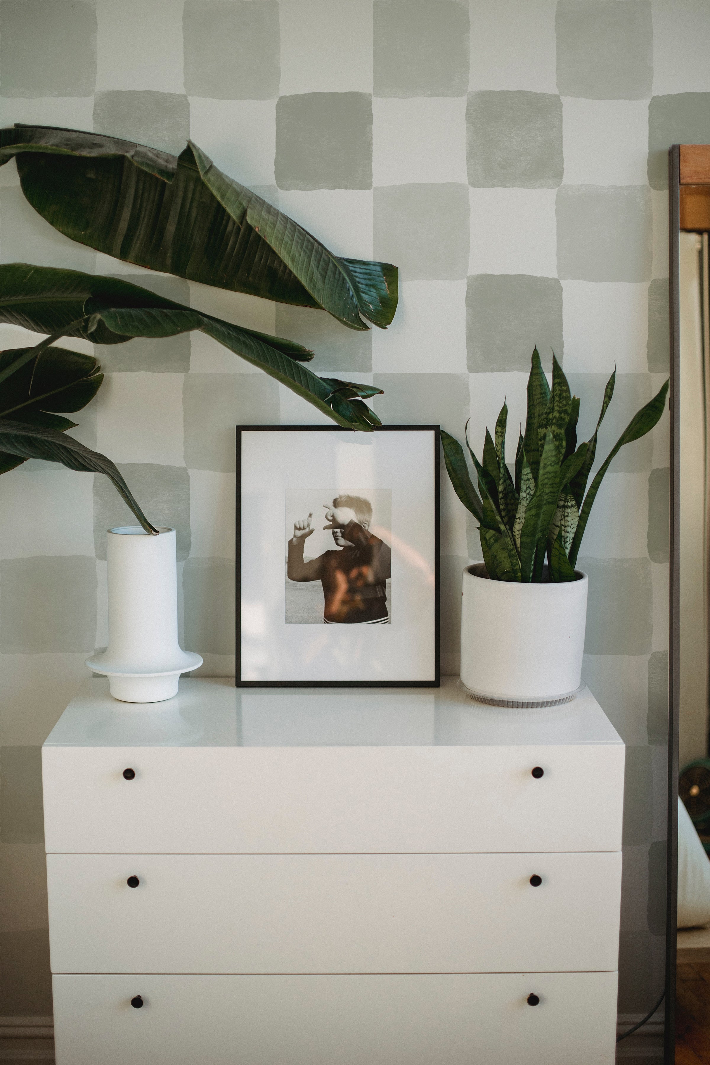 An elegant interior decorated with the Clémence Wallpaper - Olive behind a modern white dresser. A large potted plant and a framed photo complement the wallpaper's hand-painted checkered pattern, creating a chic and natural ambiance.