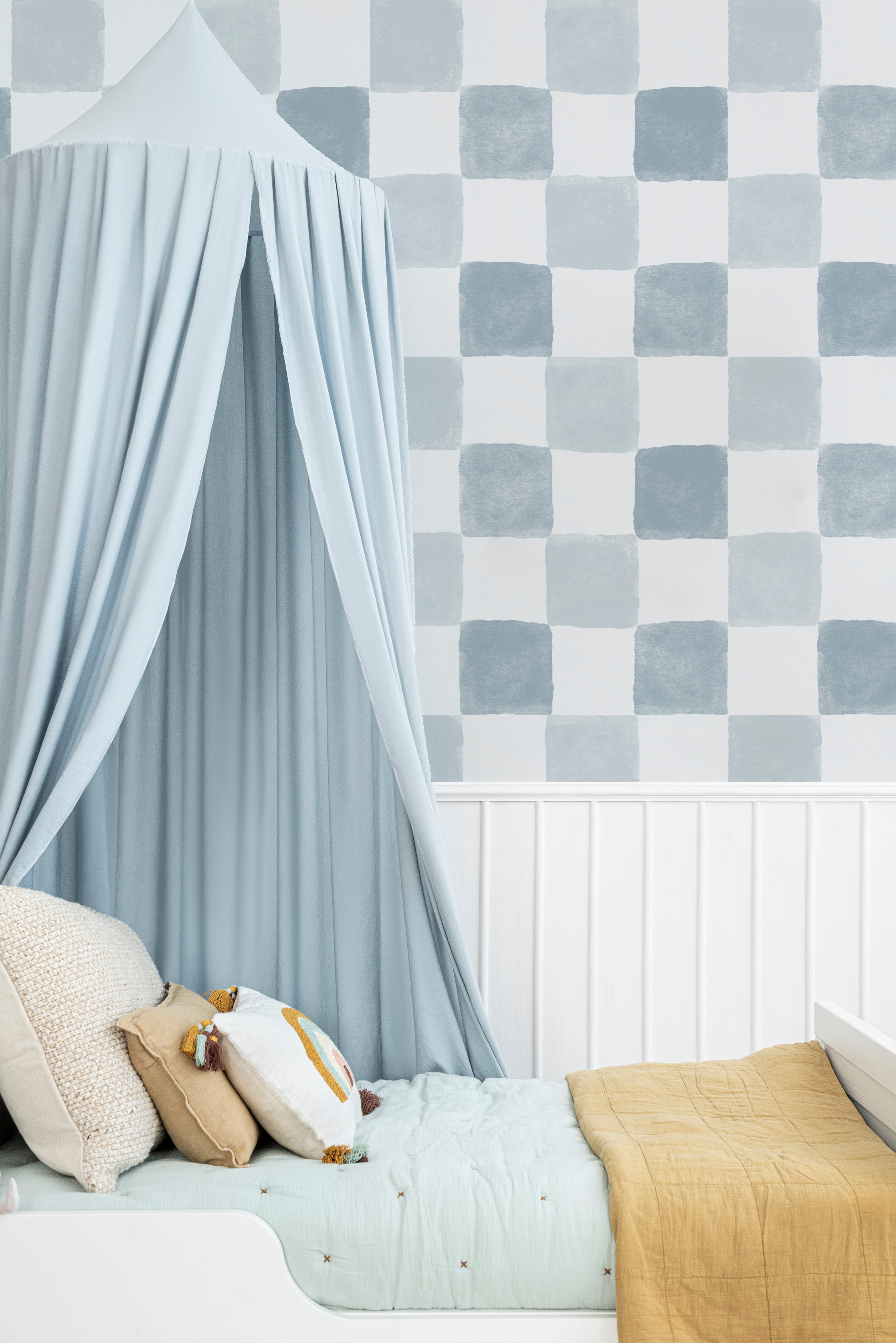 A cozy children's nook featuring Clémence Wallpaper in Pale Blue, displaying a checkered pattern with alternating soft pale blue and white squares. The space is accented with a whimsical blue canopy draped over a bed, complemented by plush pillows and a mustard blanket.