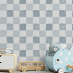 A playful child’s room adorned with Clémence Wallpaper in Pale Blue, highlighting a checkered pattern of pale blue and white squares. The room includes a white wooden toy storage unit, colorful round mats, and various playful toys, creating a charming and fun space