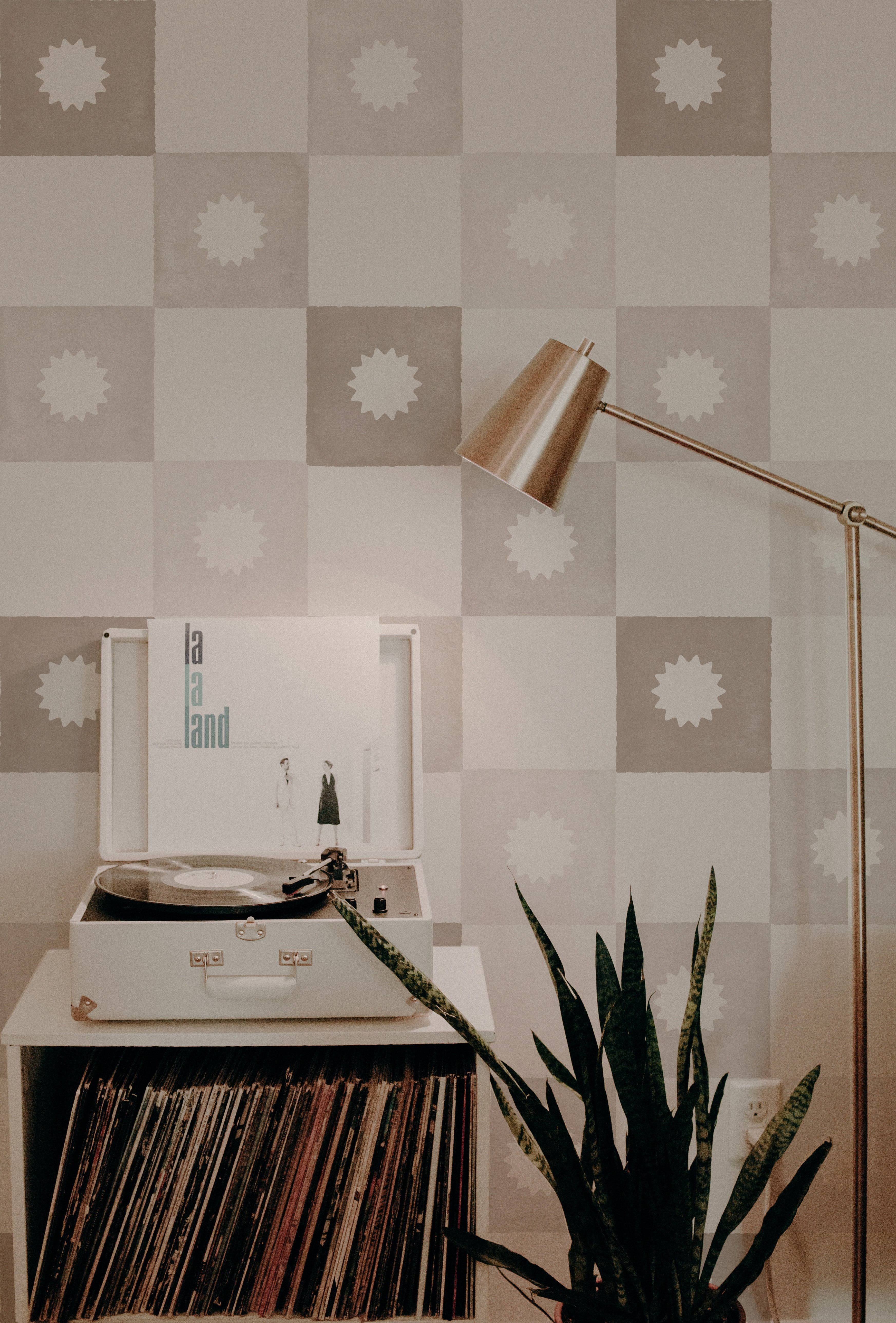 A cozy corner featuring a record player setup against a beige and white checkered wallpaper. The pattern includes a unique white floral emblem in each beige square. A sleek floor lamp, a snake plant, and a collection of vinyl records complete the retro-modern vibe.