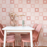 A bright and inviting dining area, decorated with Coralie Wallpaper in Dusty Pink, displaying a checkered pattern with pink starburst designs. The space is styled with pink chairs and a white table, enhancing the room’s cheerful and warm aesthetic.