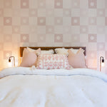 A serene bedroom featuring a large, plush bed with multiple pillows against a soft, pink and white checkered wallpaper with a subtle floral emblem in each square. Two stylish black wall-mounted lamps flank the bed.