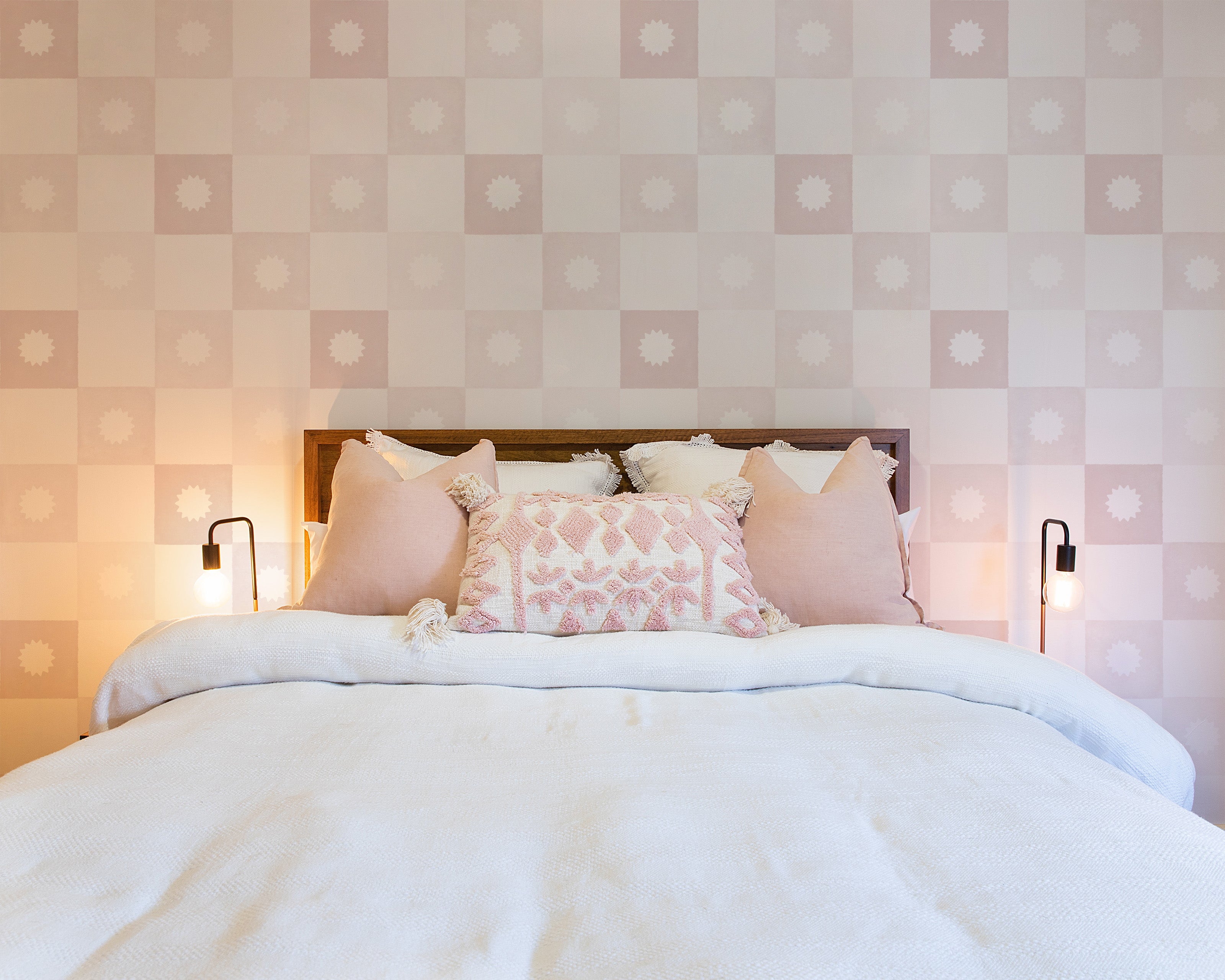 A serene bedroom featuring a large, plush bed with multiple pillows against a soft, pink and white checkered wallpaper with a subtle floral emblem in each square. Two stylish black wall-mounted lamps flank the bed.