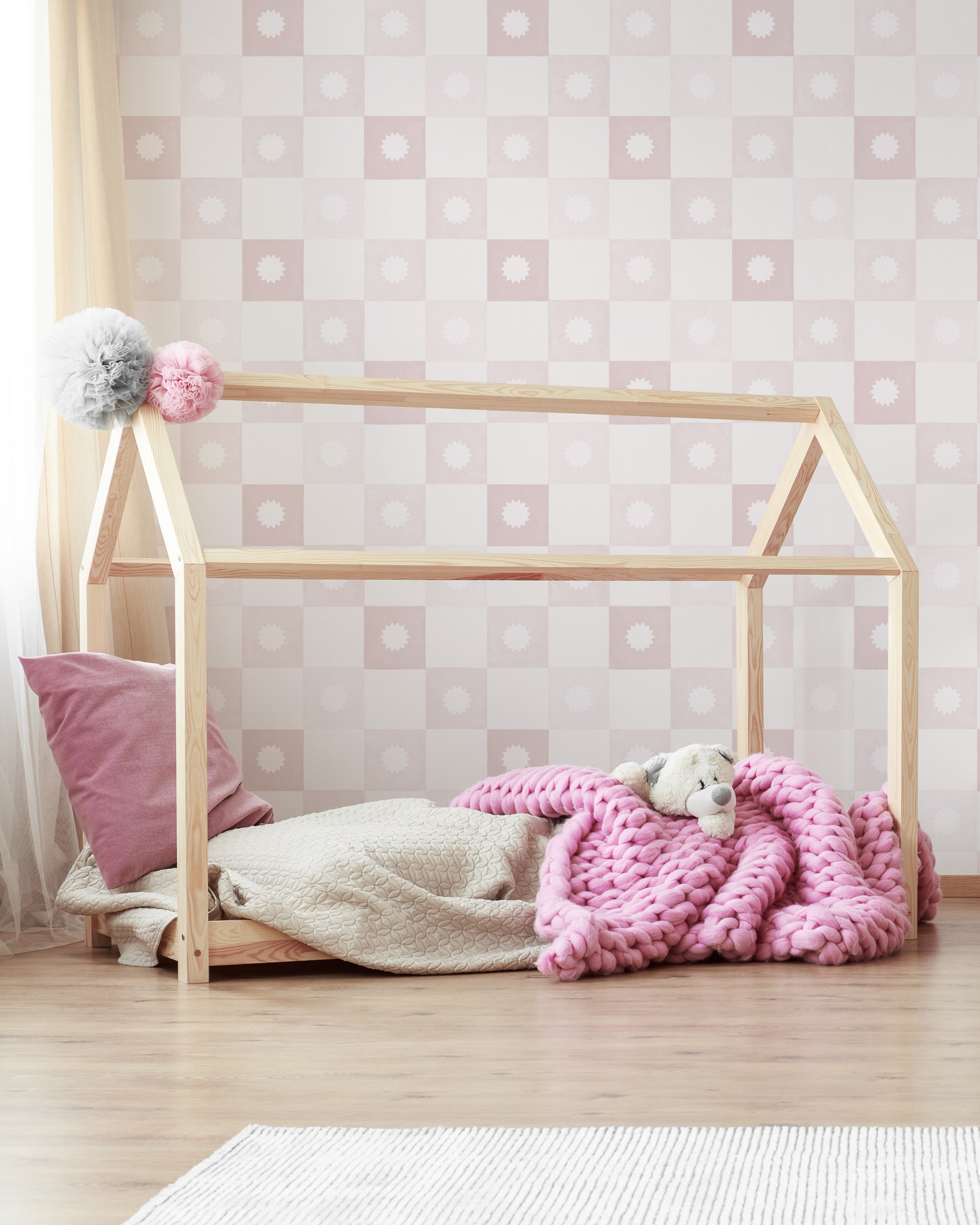 A charming children's room with a wooden frame bed set against a backdrop of pink and white checkered wallpaper. The room is decorated with soft pink cushions, a thick pink knit blanket, and stuffed toys, creating a cozy and inviting space.
