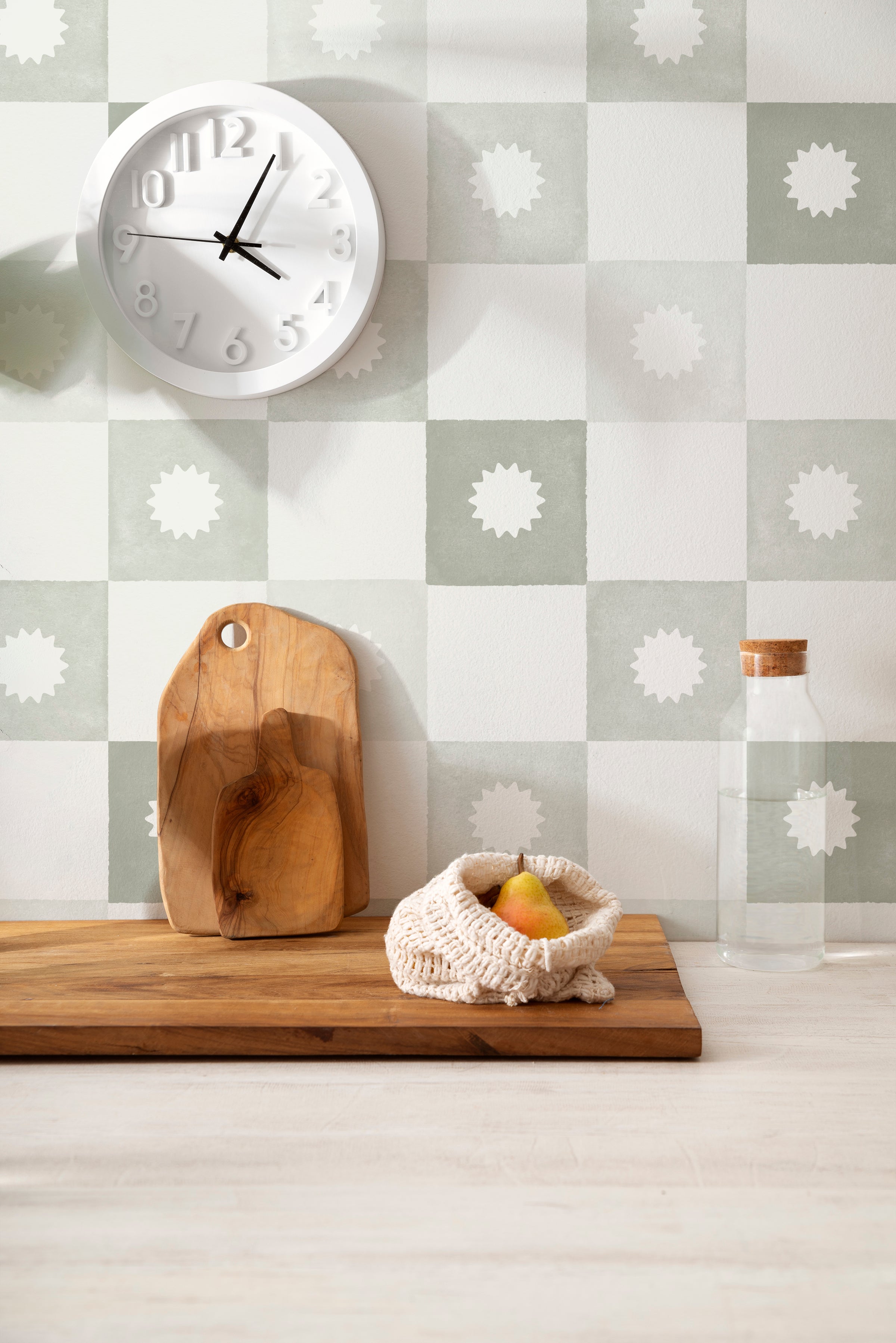 A modern kitchen setup against a wall adorned with olive and white checkered wallpaper featuring a distinct white floral emblem in each olive square. A white wall clock, a rustic wooden chopping board, and a water bottle add to the functional yet stylish decor.