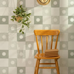 A cozy corner featuring a wooden chair against an olive and white checkered wallpaper background. A hanging potted plant and a straw hat add a touch of rustic charm, emphasizing the wallpaper's gentle and inviting design.