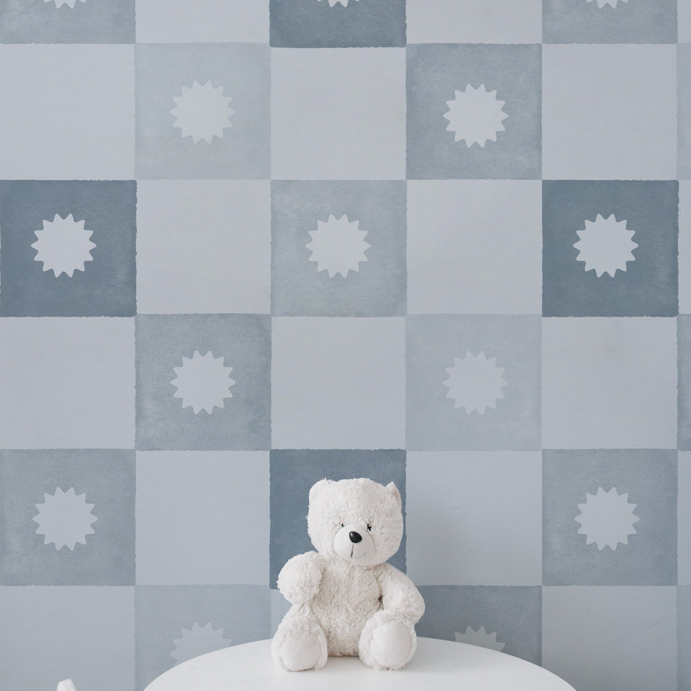 A nursery setting showing the Coralie Wallpaper in pale blue on the wall, decorated with a checkered pattern of pale blue and white squares, each with a centered sunburst motif. The scene includes a white teddy bear sitting on a white round table