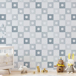 A nursery room featuring the Coralie Wallpaper in pale blue with a checkered pattern that includes alternating white and pale blue squares with a watercolor-style sunburst motif. The room includes a white crib, a small white table, plush toys, and a circular grey rug on a wooden floor.
