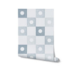 A roll of Coralie Wallpaper in pale blue, unrolled partially to reveal the checkered pattern. Each square alternates between pale blue and white, each with a centered sunburst motif, depicted in a soft watercolor style.