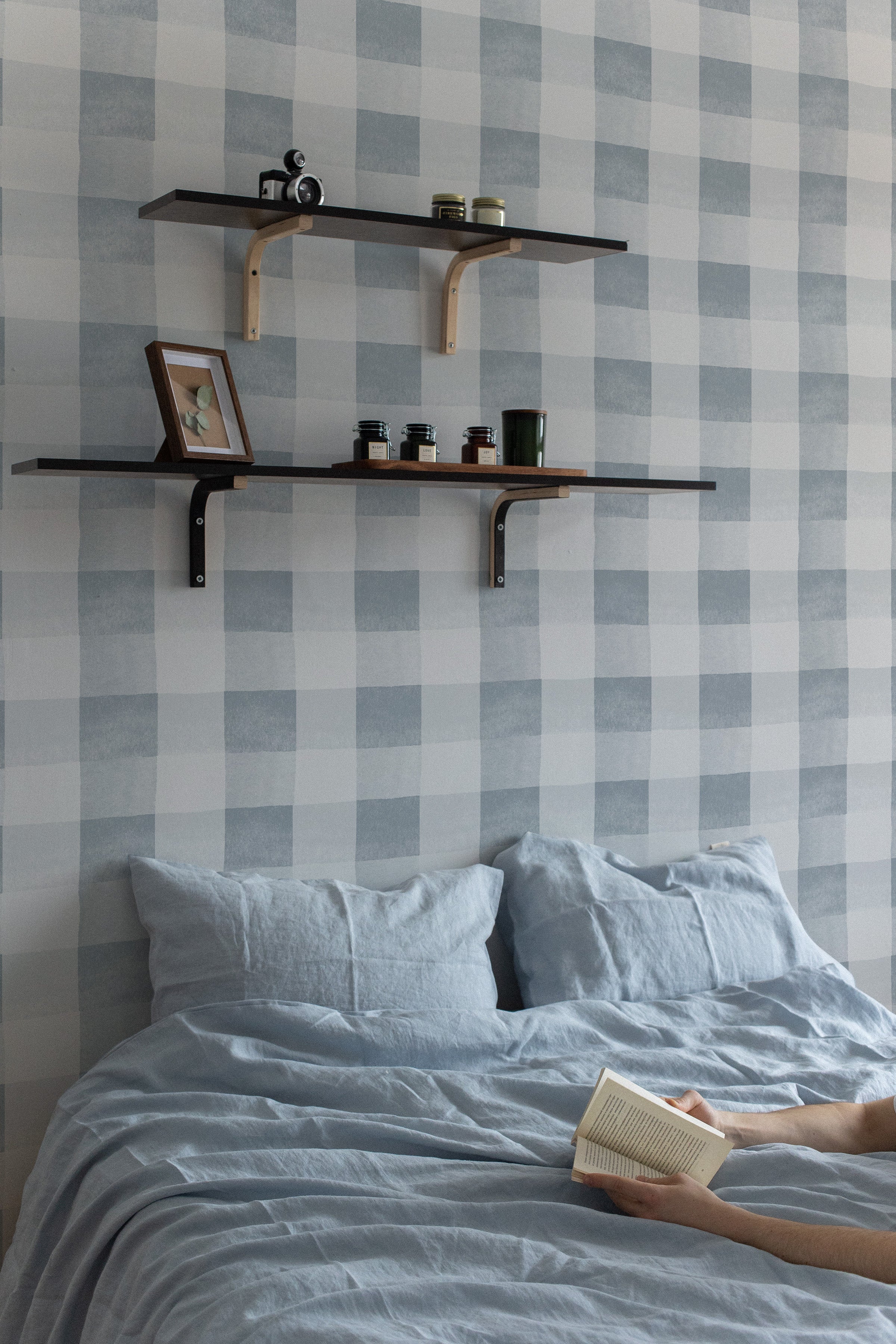  a bedroom interior with "Camille Wallpaper - Pale Blue" adorning the wall behind a cozy bed with blue linens. The gentle pale blue checkered pattern of the wallpaper provides a serene backdrop, enhancing the room's relaxed vibe. Wooden shelves with personal items and plants add warmth to the space, complementing the soft aesthetic of the wallpaper.