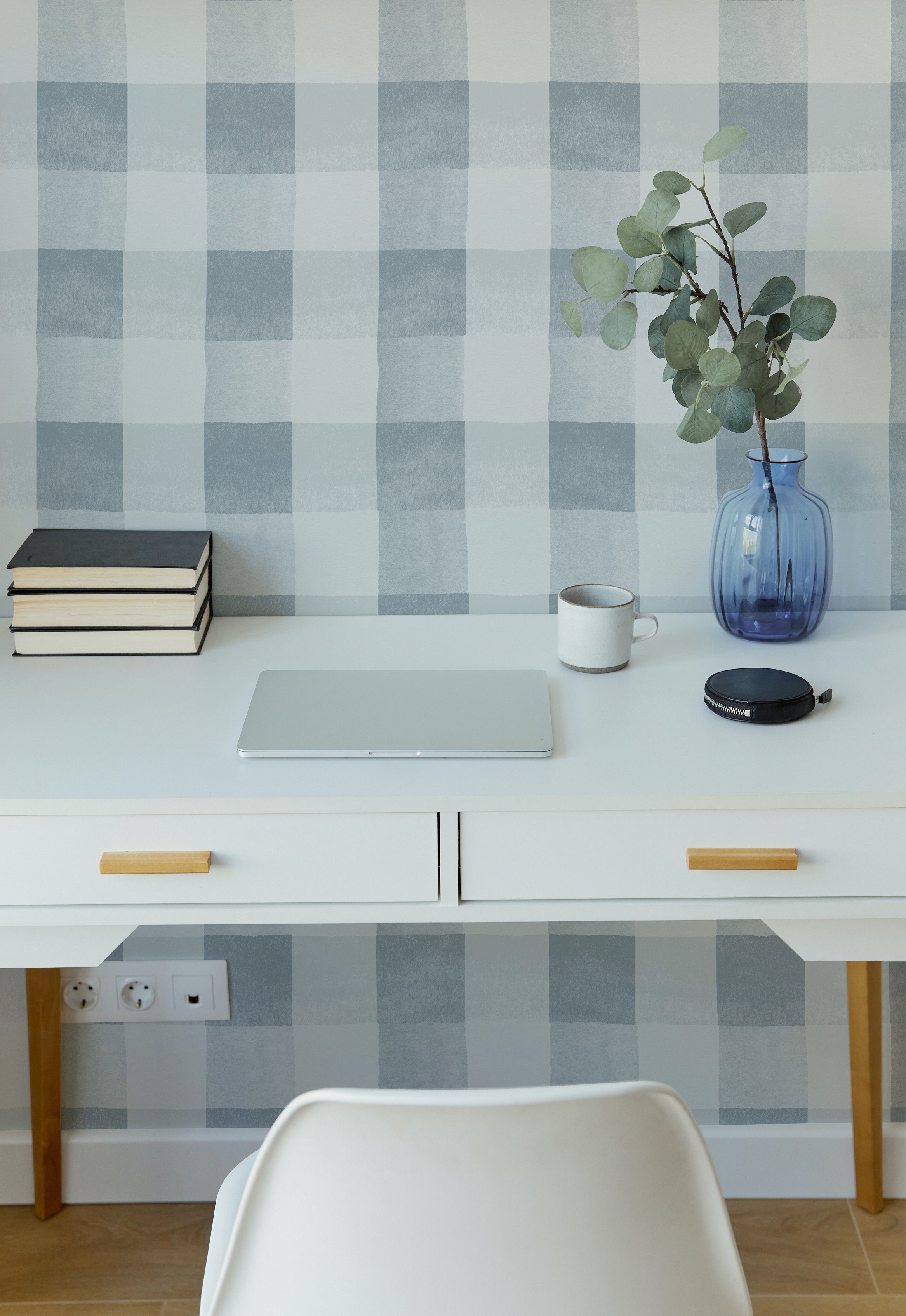 a modern home office setting with "Camille Wallpaper - Pale Blue" on the wall. The wallpaper displays a subtle, pale blue and white checkered pattern. The room features a sleek white desk with wooden handles, a laptop, a stack of books, a cup, and a stylish round storage box. A white chair is placed in front of the desk, and a vibrant blue vase with eucalyptus branches adds a touch of color to the serene palette.
