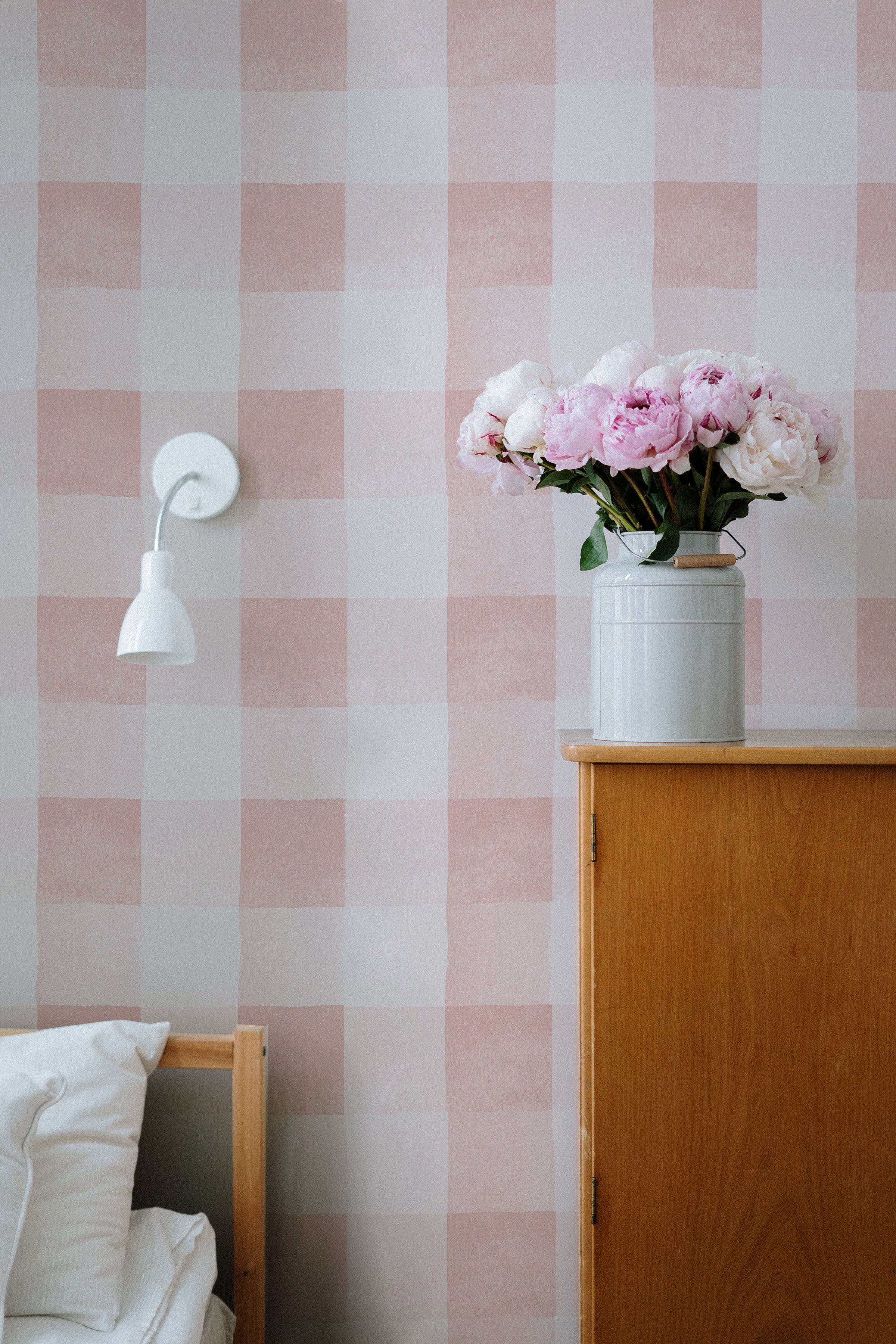 A serene bedroom corner featuring Camille Wallpaper in dusty pink with a large checkered pattern. The soft pink tones of the wallpaper provide a warm and comforting ambiance, complemented by a white bedside lamp and a vase with fresh pink peonies on a wooden cabinet