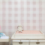 A serene nursery setting with Camille Wallpaper - Nude, featuring a soft checkered pattern in shades of pale pink and white. The gentle backdrop complements the vintage white dresser used as a changing table, adorned with a pink lamp and a plush bunny, creating a warm and inviting space for a child