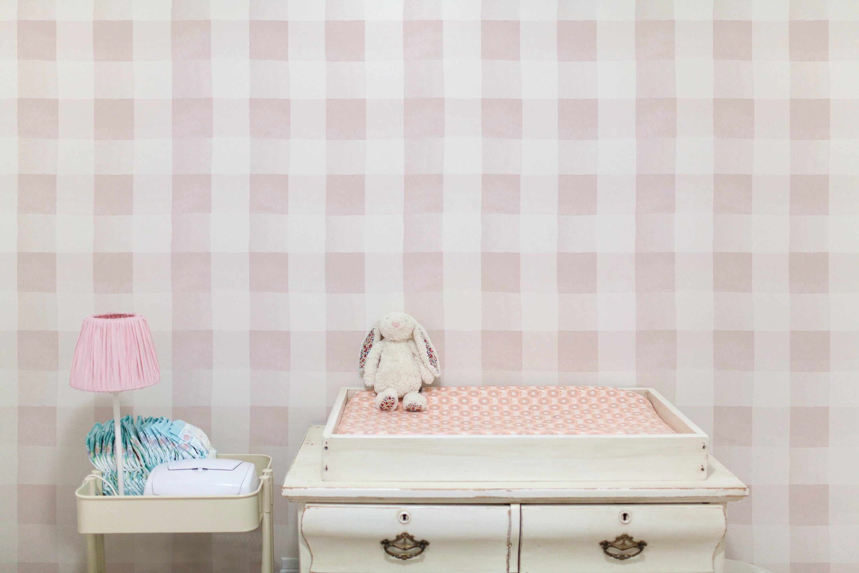 A serene nursery setting with Camille Wallpaper - Nude, featuring a soft checkered pattern in shades of pale pink and white. The gentle backdrop complements the vintage white dresser used as a changing table, adorned with a pink lamp and a plush bunny, creating a warm and inviting space for a child