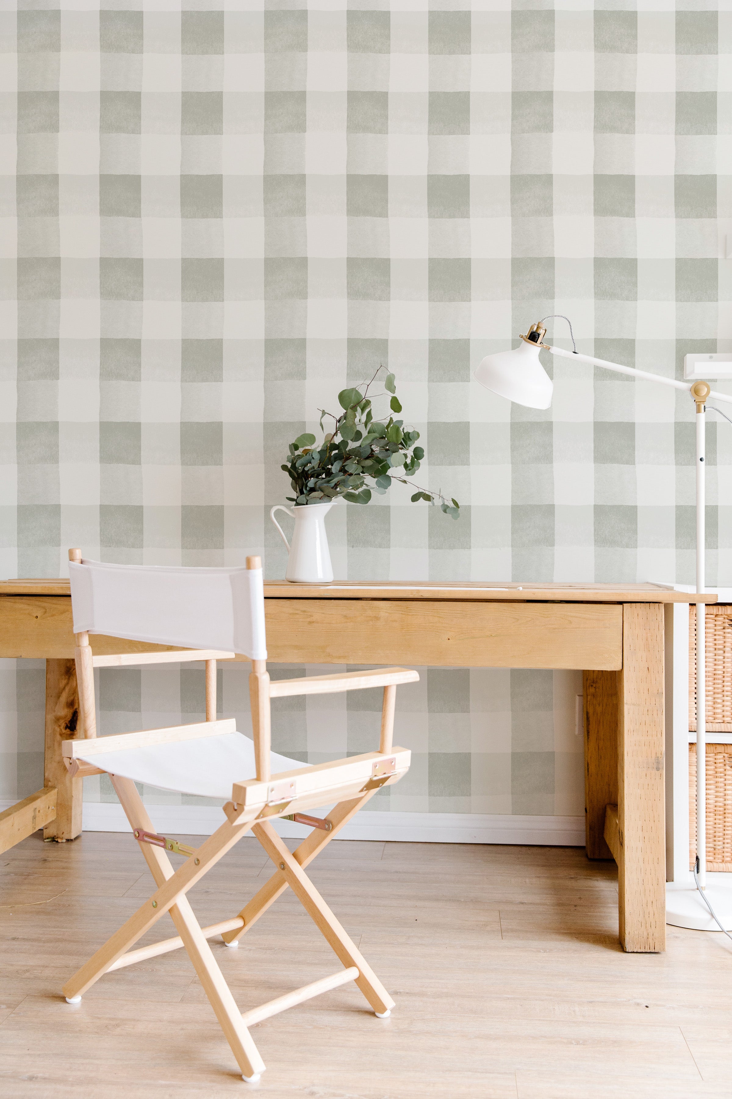 A vertical image featuring the Camille Wallpaper - Olive in a modern workspace. The wallpaper displays a large, olive green checkered pattern on an off-white background. A wooden desk with a director's chair in white, a vase with eucalyptus branches, and a white desk lamp complete this simple yet stylish scene.