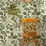 an interior scene with "Green Goddess Wallpaper." A single wooden chair with a woven hat hanging above it is set against the wallpaper, highlighting the room's natural aesthetic. A potted hanging plant with greenery that complements the wallpaper adds a three-dimensional element to the space.