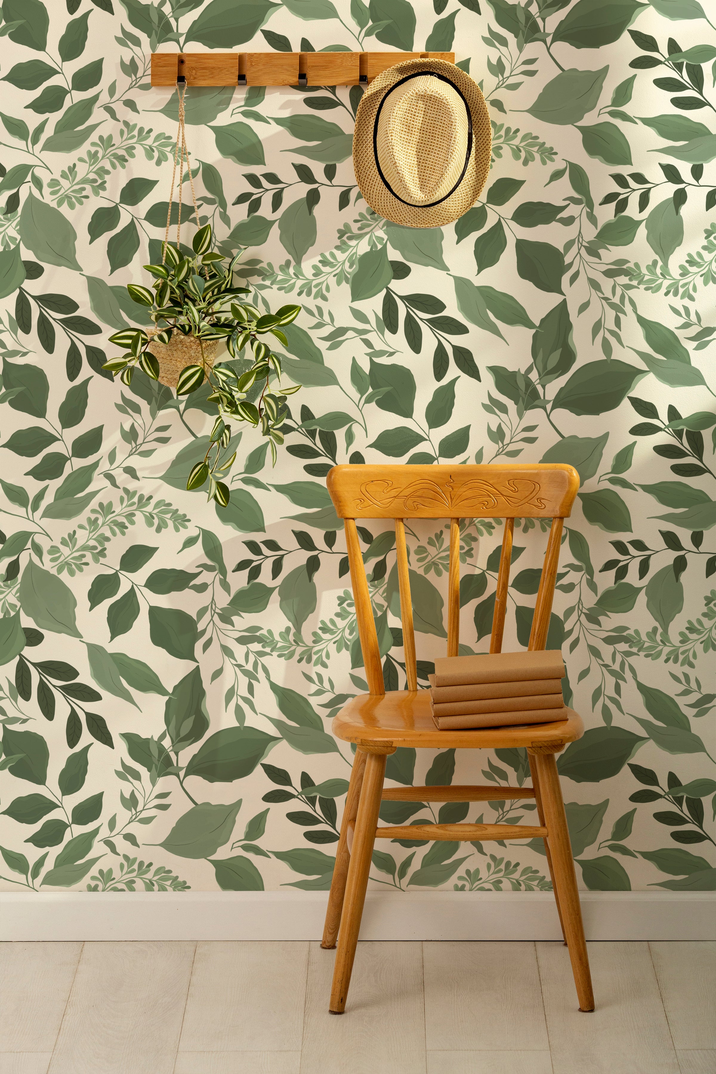 an interior scene with "Green Goddess Wallpaper." A single wooden chair with a woven hat hanging above it is set against the wallpaper, highlighting the room's natural aesthetic. A potted hanging plant with greenery that complements the wallpaper adds a three-dimensional element to the space.