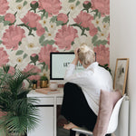 A home office space elegantly decorated with Maggie Floral Wallpaper, displaying an array of pink peonies and white flowers. The vibrant floral pattern creates a lively and inspiring workspace, complemented by a white desk, a laptop, and green indoor plants.