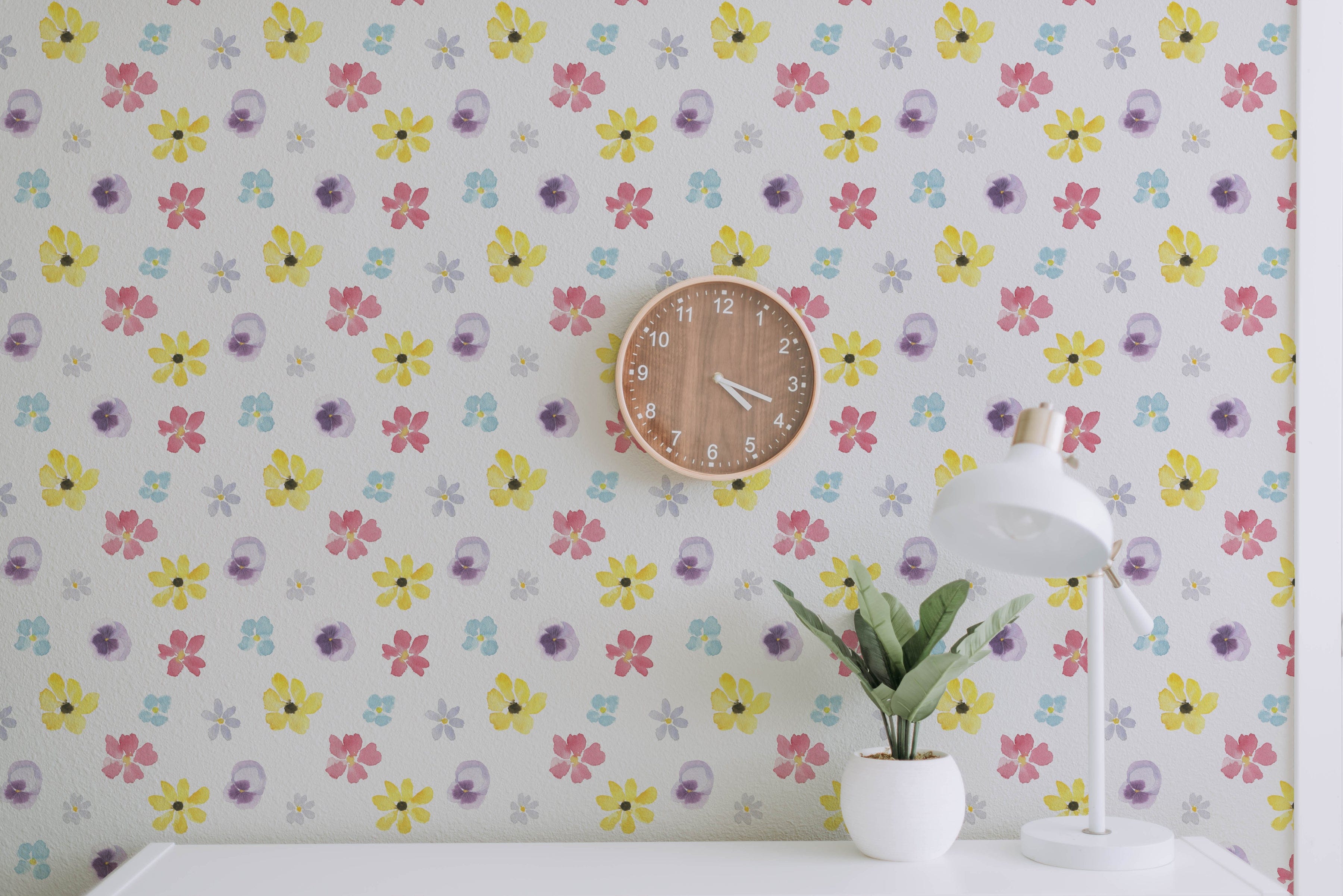 A workspace adorned with the Spring Field Wallpaper - VI, highlighting its lively floral pattern in bright yellow, pink, purple, and blue hues. The space includes a wooden wall clock and a white desk lamp beside a small plant.