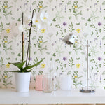 A stylish room corner with the Spring Field Wallpaper - VII, featuring a white orchid in a pot and a sleek metallic desk lamp on a white desk. The wallpaper's design includes purple and yellow flowers and green foliage