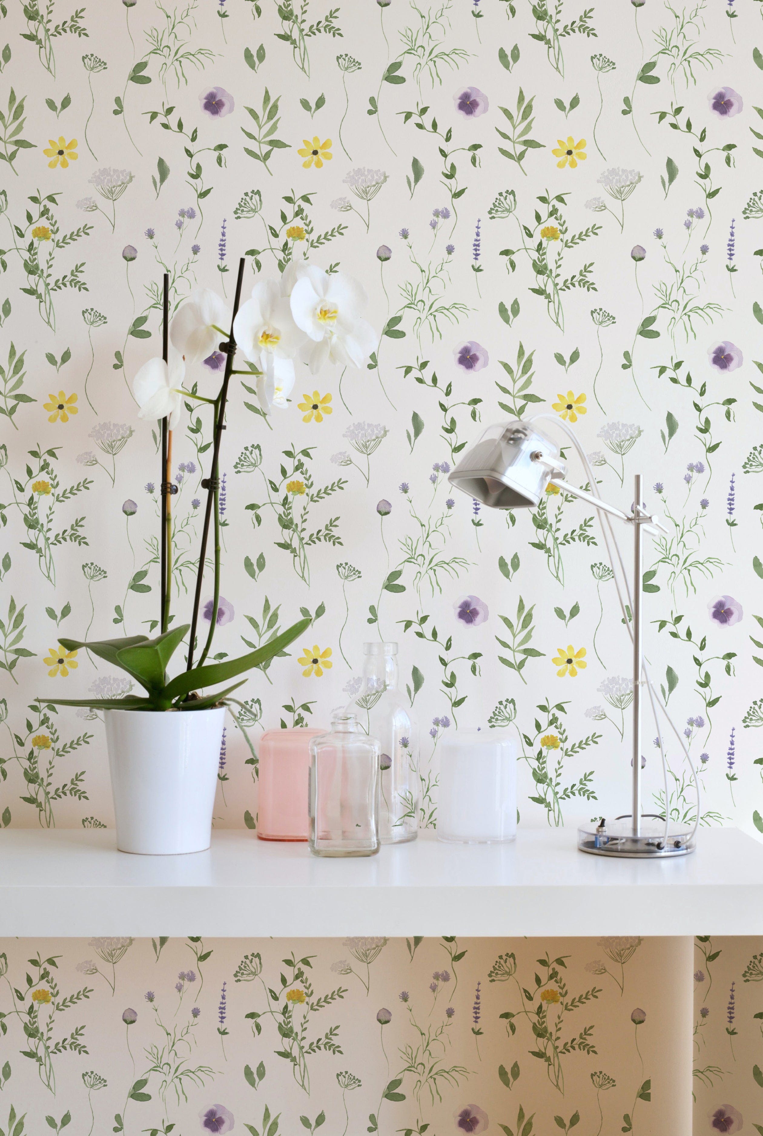 A stylish room corner with the Spring Field Wallpaper - VII, featuring a white orchid in a pot and a sleek metallic desk lamp on a white desk. The wallpaper's design includes purple and yellow flowers and green foliage