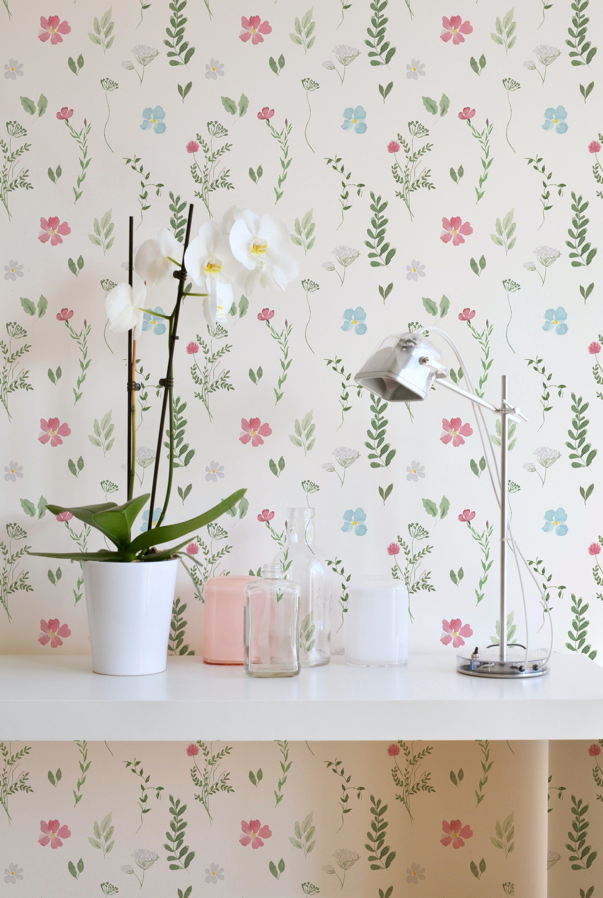 A stylish room corner with the Spring Field Wallpaper - VIII, highlighting its elegant floral pattern in pink, blue, and yellow hues. The decor includes a white orchid in a pot and a sleek metallic desk lamp on a white desk