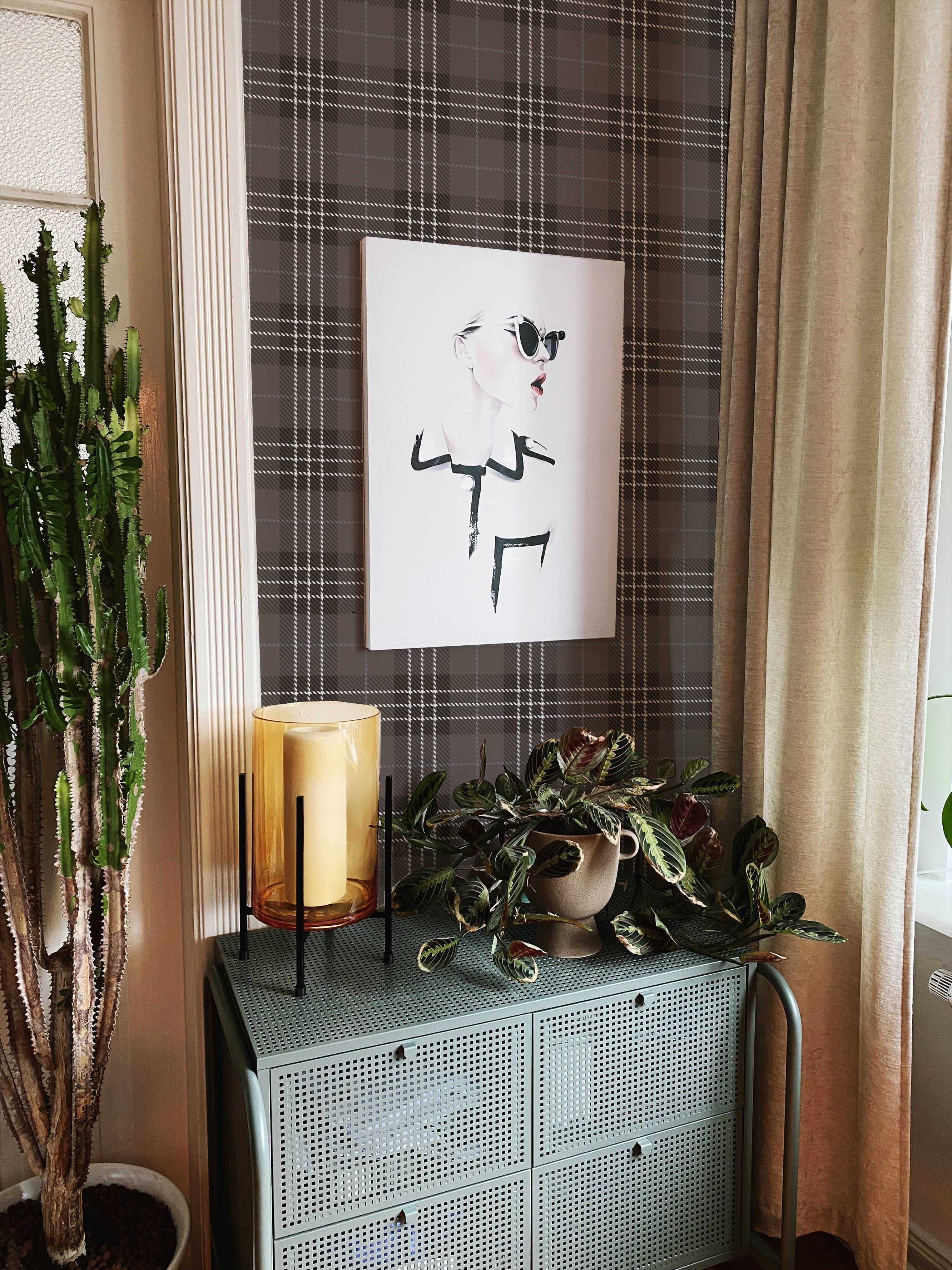 A snippet of home decor featuring the Dark Plaid Wallpaper - Black behind an art piece, flanked by indoor plants and a large candle, depicting an intimate corner that blends classic patterns with contemporary design elements for a unique, stylish ambiance.