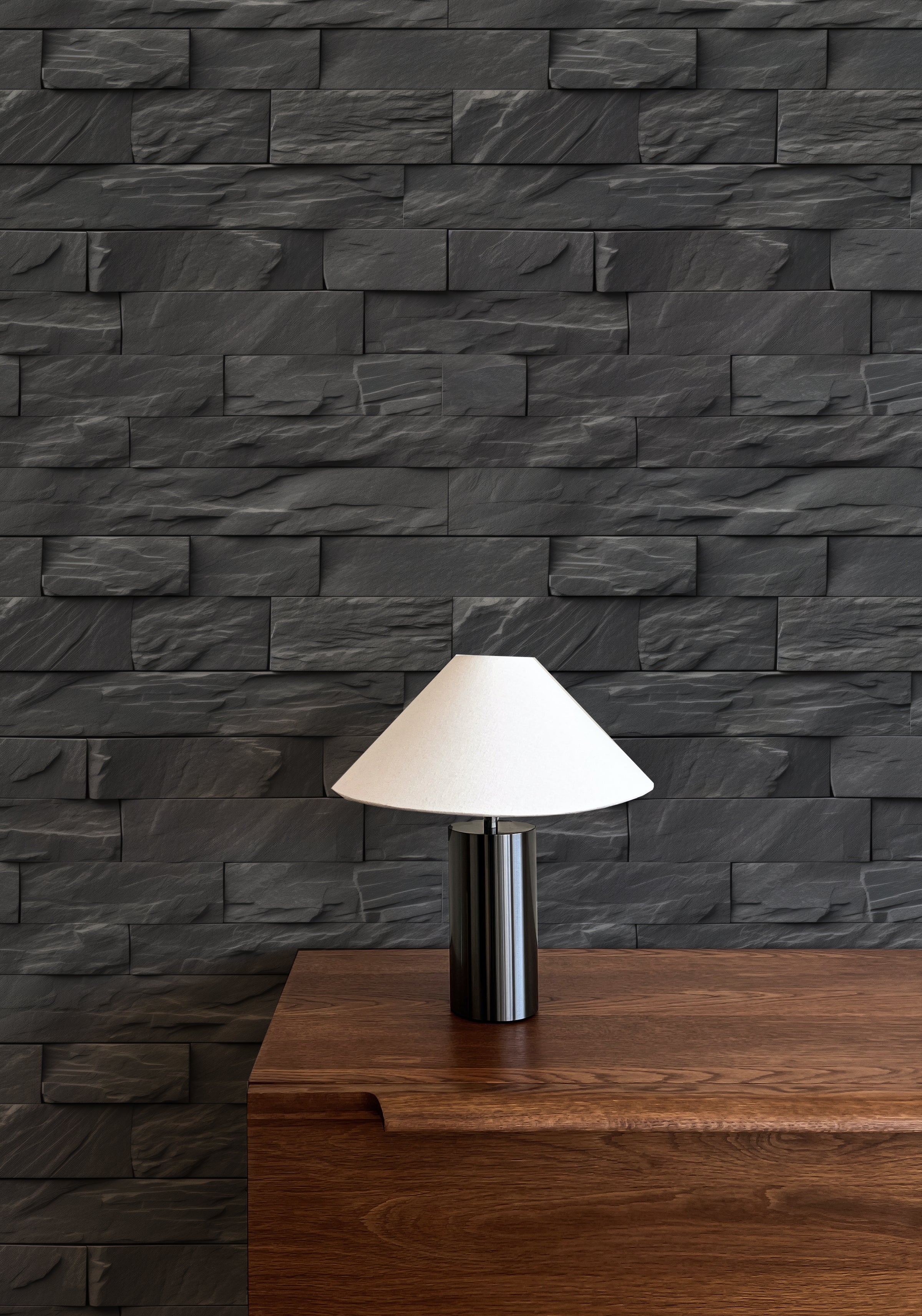 A modern interior with Black Brick Wallpaper featuring a textured, stacked stone design in dark gray. The wallpaper provides a striking backdrop for a wooden sideboard and a minimalist black table lamp with a white shade.