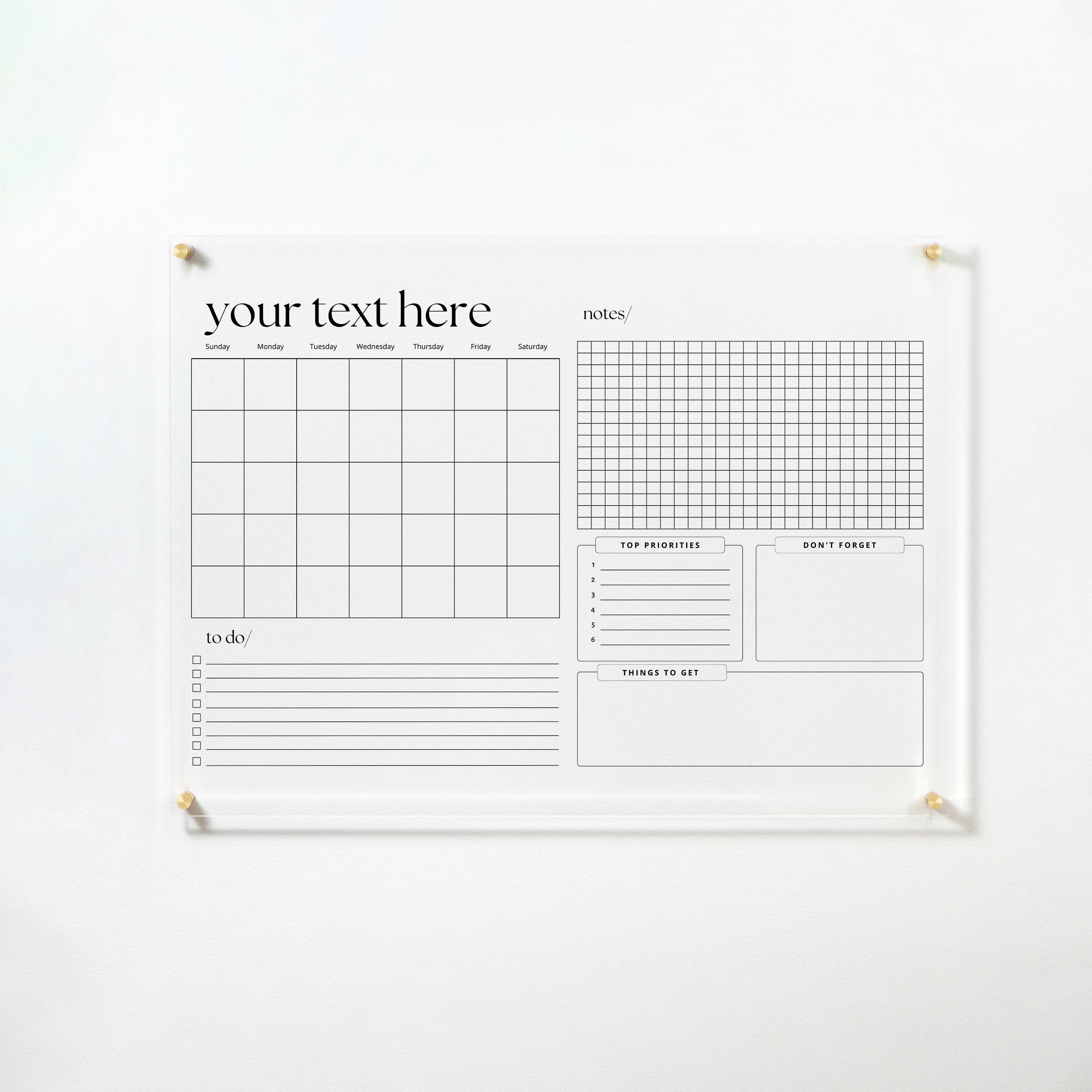 The full view of the same acrylic board, mounted on a white wall with gold pins. The board includes a monthly calendar, to-do list with checkboxes, a grid notes section, and areas for top priorities, things to get, and don't forget, all in a sleek black and white design.