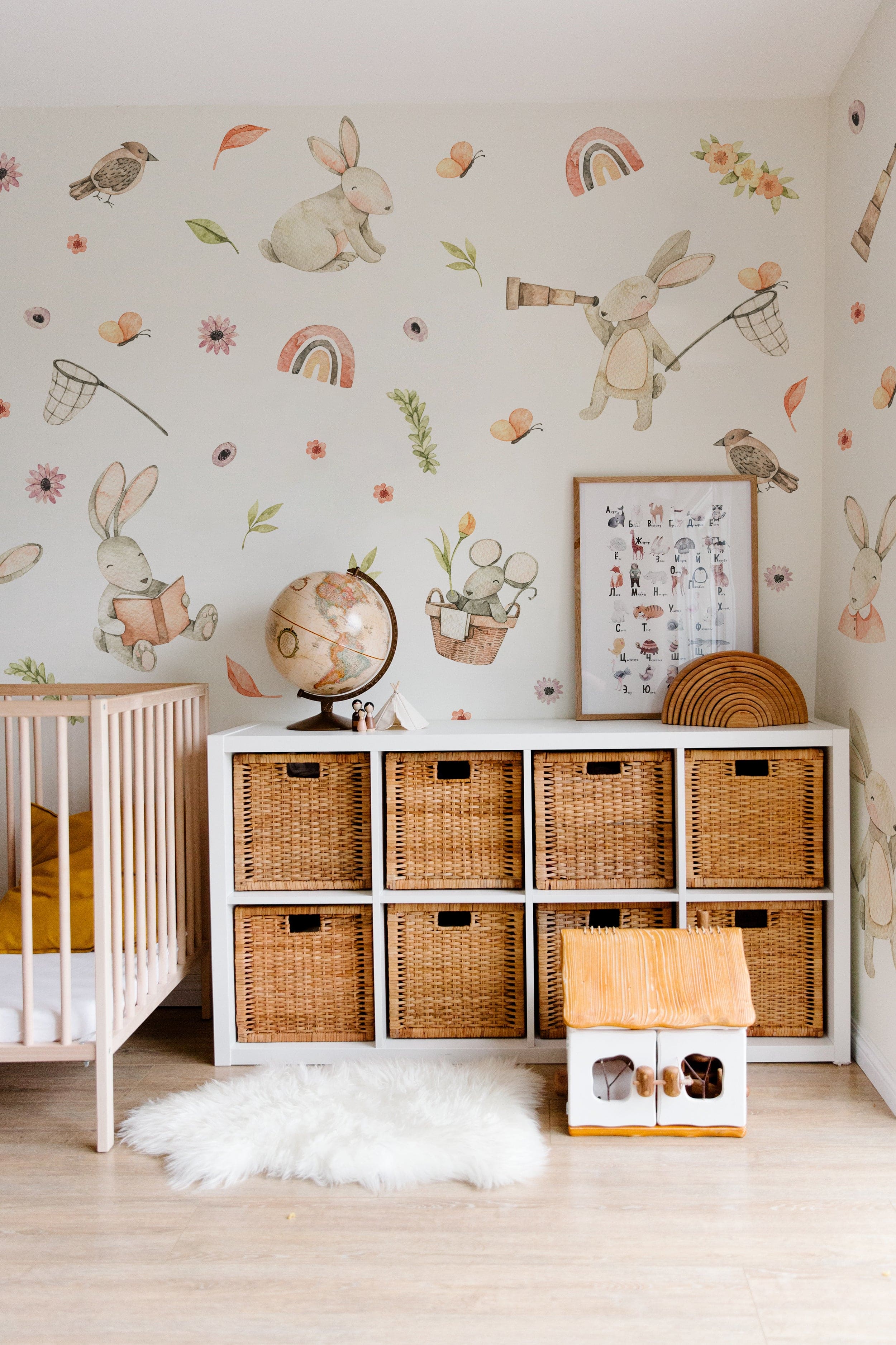 Children's room with watercolour bunny wall decals featuring playful bunnies, birds, butterflies, flowers, and rainbows on a white wall above a storage unit with baskets.