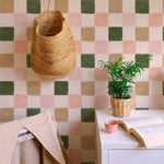 A cozy corner with Clémence Wallpaper II on the wall, showing the checkered pattern in green, beige, pink, and white. A wicker basket hangs on the wall, and a small potted plant sits on a white desk with an open book and a pink cup.