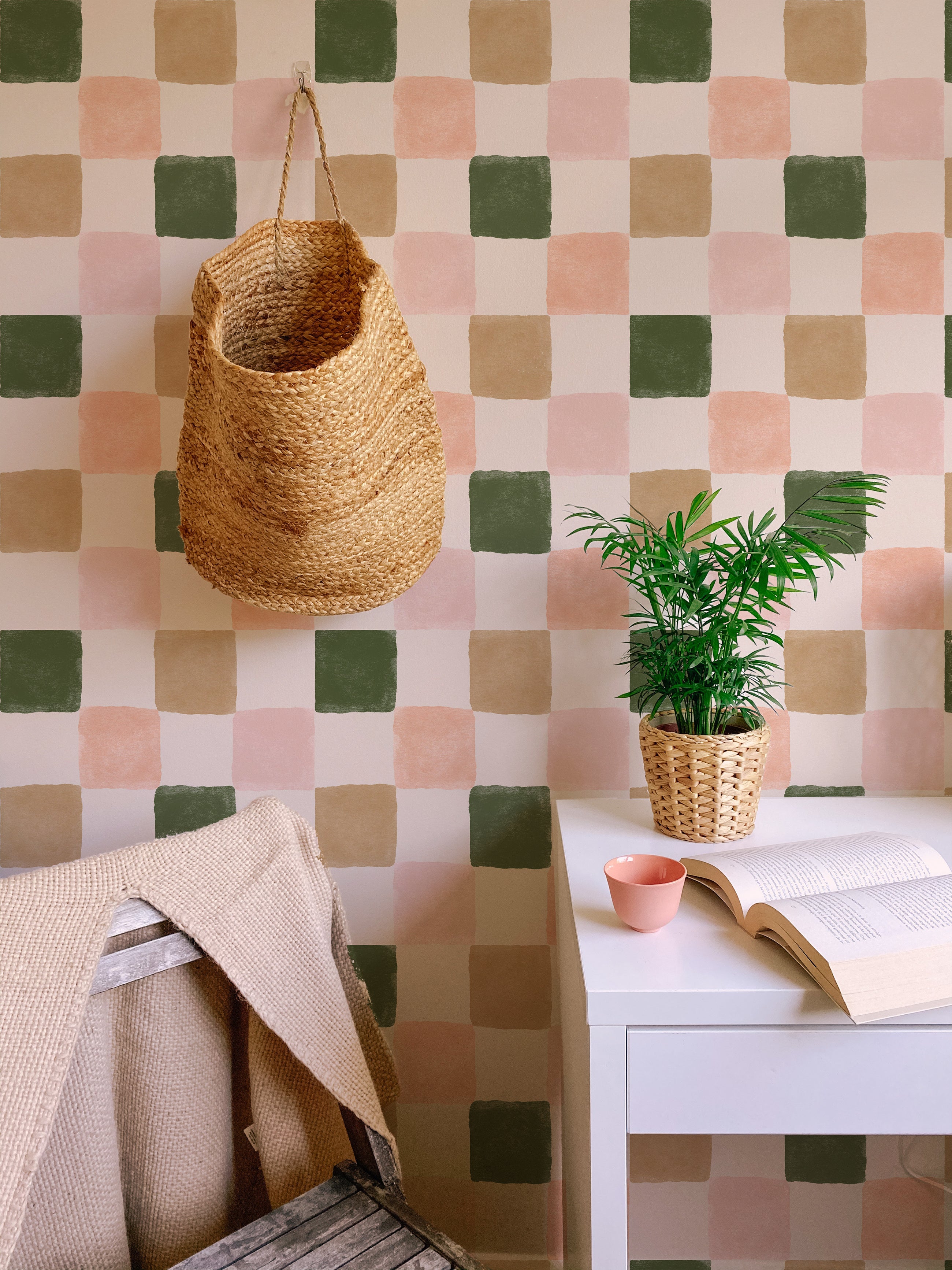 A cozy corner with Clémence Wallpaper II on the wall, showing the checkered pattern in green, beige, pink, and white. A wicker basket hangs on the wall, and a small potted plant sits on a white desk with an open book and a pink cup.