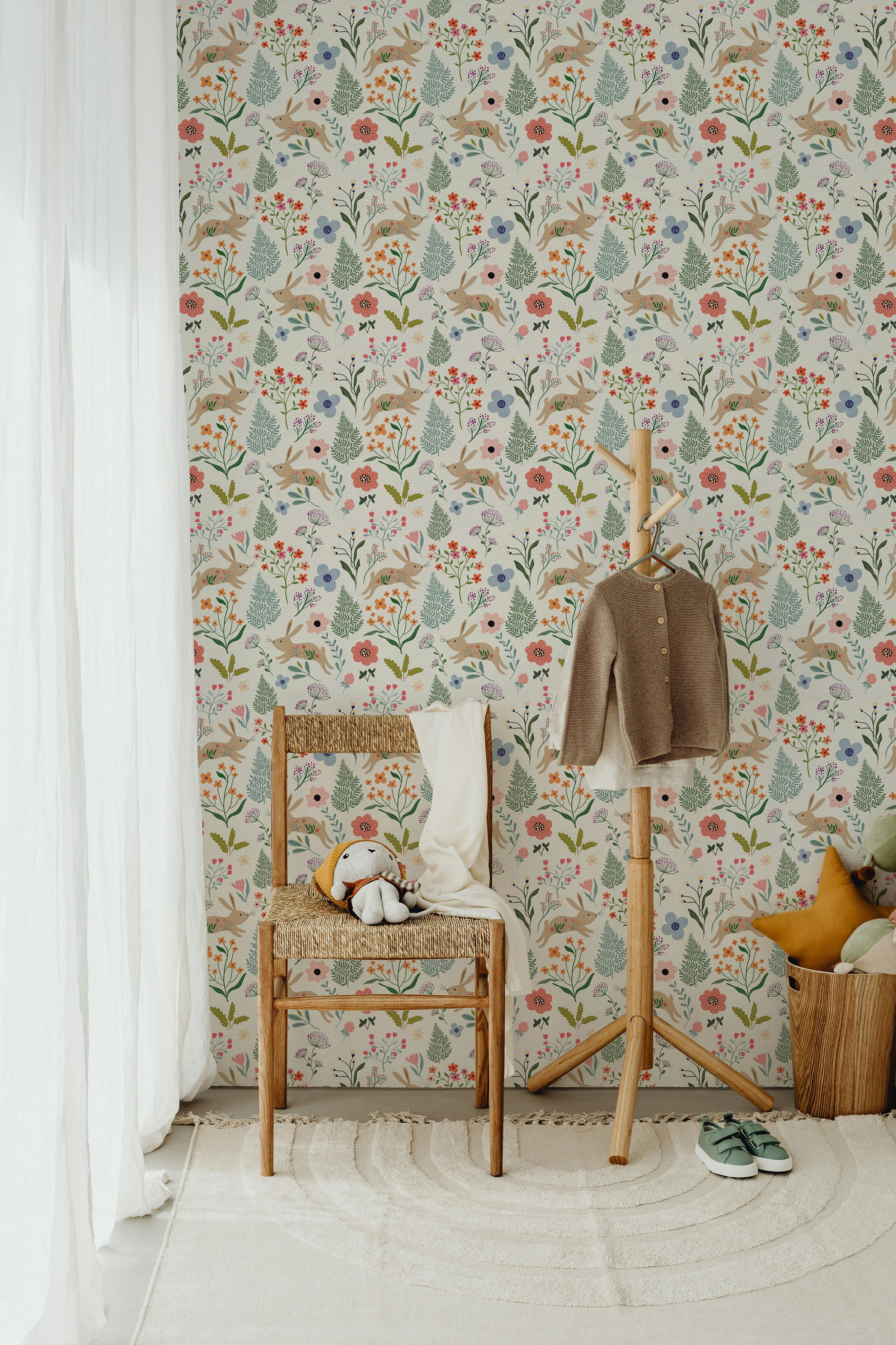 A cozy corner of a room decorated with the Colourful Spring Bunnies Wallpaper, accompanied by a rustic wooden chair and a small white curtain. The wallpaper's mix of bunnies and assorted floral patterns creates a peaceful, nature-inspired ambiance.
