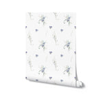 A roll of Delicate Blue Wildflower Wallpaper reveals its charming and detailed botanical print. The light background adorned with delicate blue flowers and soft green leaves offers a fresh and inviting look for a room update