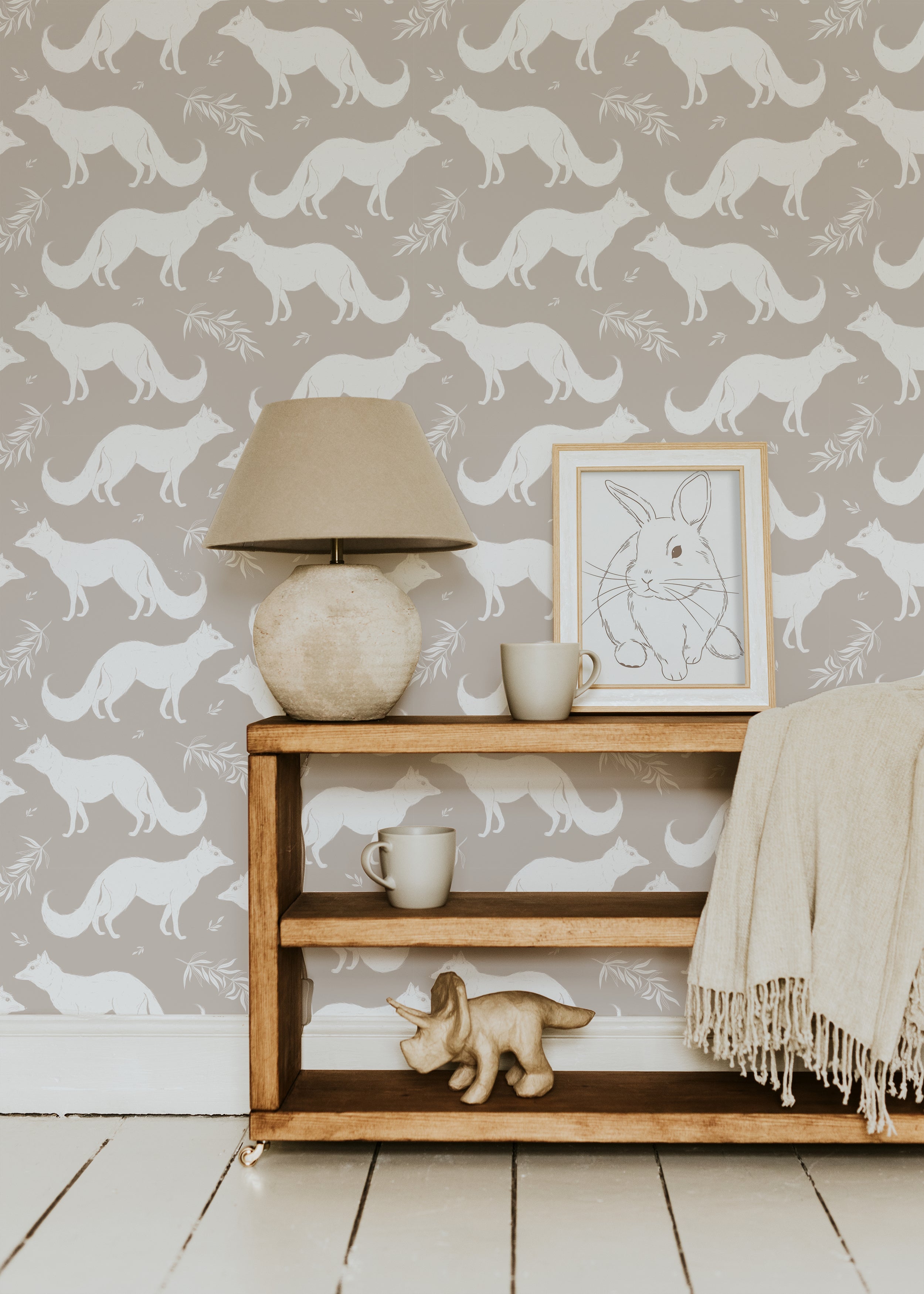 A cozy living room setting with a wooden shelf, ceramic lamp, and framed rabbit illustration in front of a warm beige wallpaper featuring white foxes and delicate foliage patterns, creating a charming and serene ambiance.