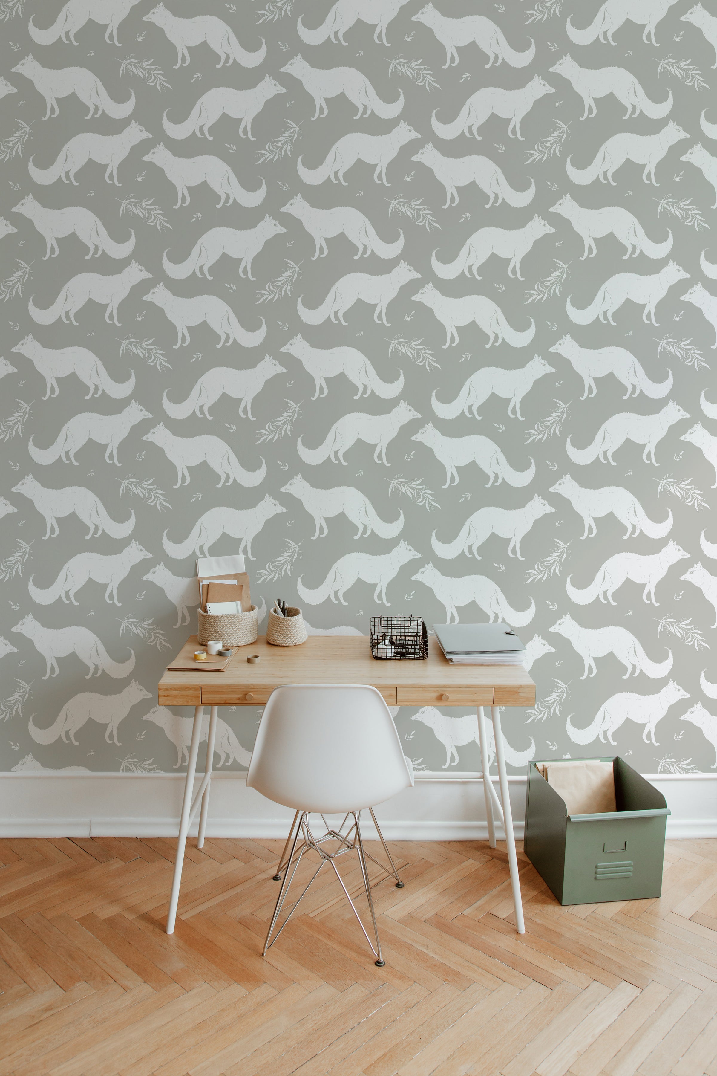 A home office with a light wooden desk and white chair placed against a muted green wallpaper adorned with a pattern of white foxes and delicate foliage, creating a calm and serene workspace