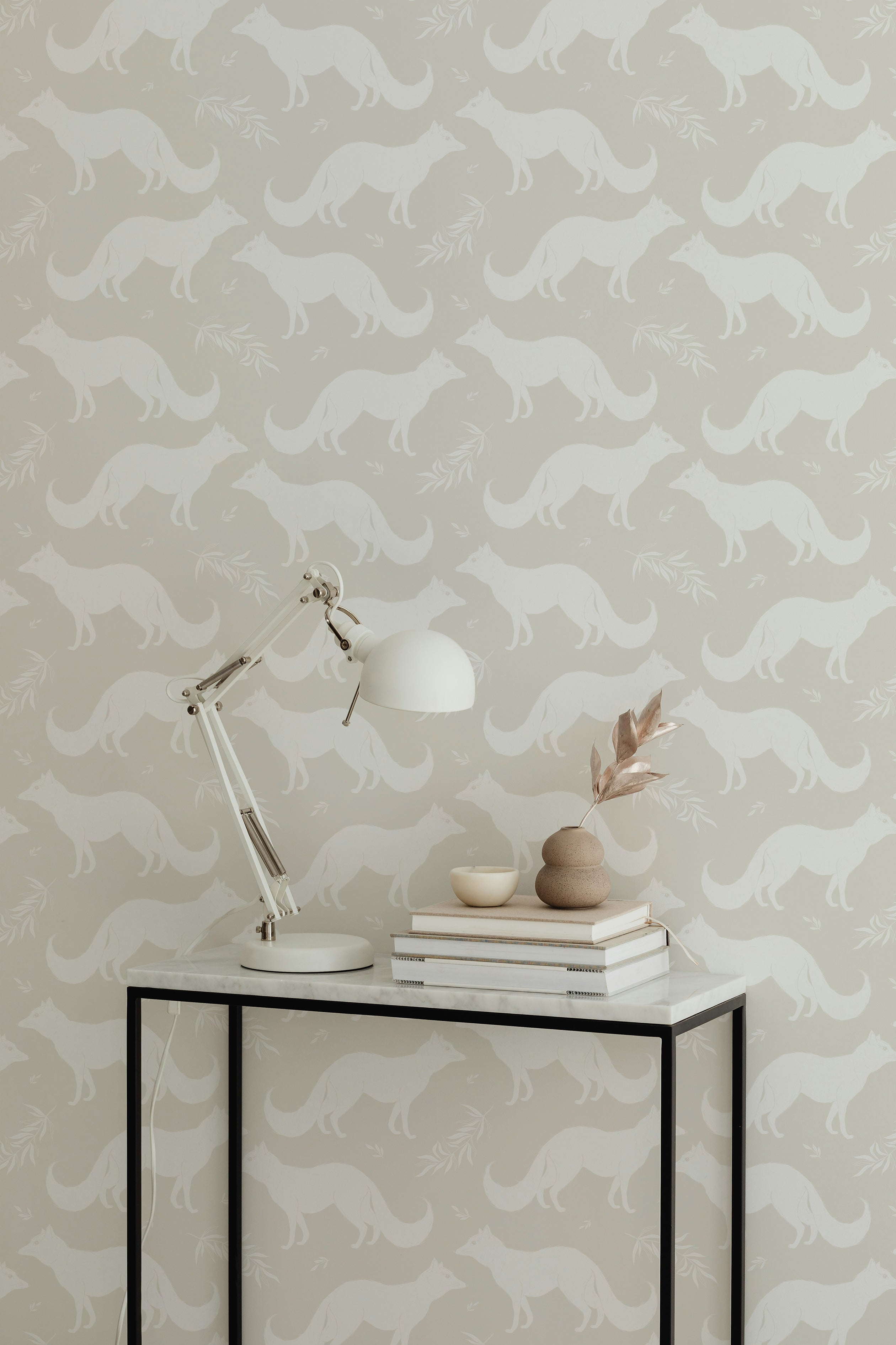 A minimalist workspace with a white desk lamp and a stack of books on a table in front of a warm beige wallpaper adorned with white foxes and delicate foliage patterns, adding a touch of nature to the room.