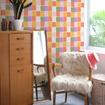 A stylish room setup showcasing the Abstract Checker Wallpaper with vibrant square tiles in alternating colors of orange, pink, purple, green, and yellow. The room features a vintage wooden cabinet, a fluffy white chair, and a large mirror, creating a cozy and modern space that highlights the wallpaper’s colorful charm.