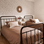 A bedroom with two single beds against a wall covered in Dainty Minimal Floral Wallpaper, exhibiting a black botanical pattern on a white backdrop, complemented by vintage metal bed frames and earth-toned bedding.