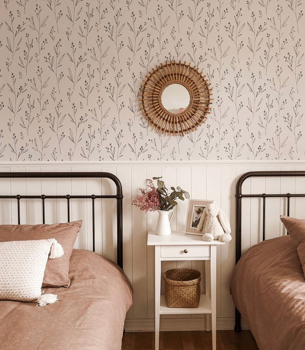 A bedroom with two single beds against a wall covered in Dainty Minimal Floral Wallpaper, exhibiting a black botanical pattern on a white backdrop, complemented by vintage metal bed frames and earth-toned bedding.