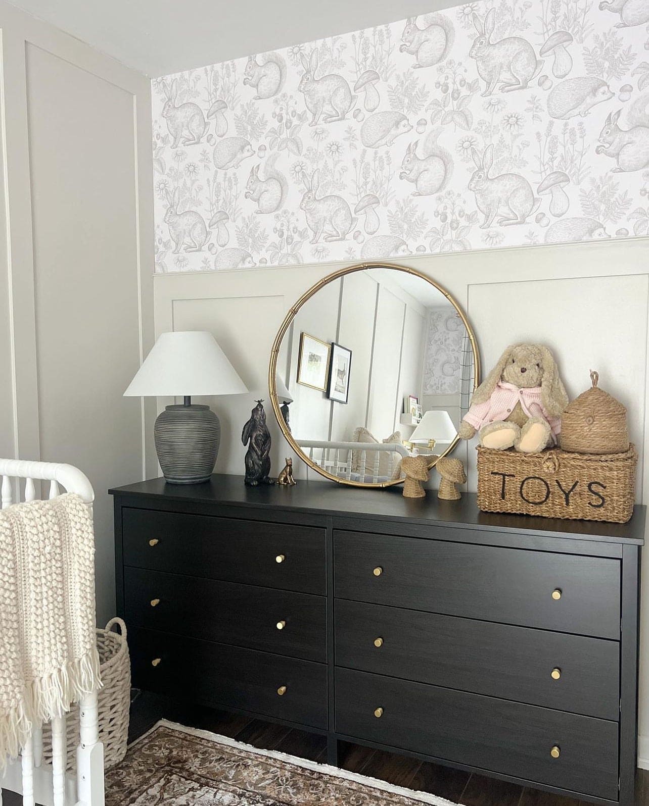 The "Woodland Creatures Wallpaper - Beige" sets a peaceful and playful backdrop in a child's room with a black dresser and a round mirror. Plush toys and a woven "TOYS" basket on the dresser match the theme, adding to the room's enchanted forest vibe.
