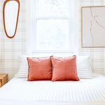 A bright bedroom setting featuring Traditional Tartan Plaid Wallpaper as the backdrop to a comfortable bed dressed in white and accented with coral pillows, the simplicity of the decor complemented by a vintage oval wooden mirror, evoking a sense of timeless elegance.