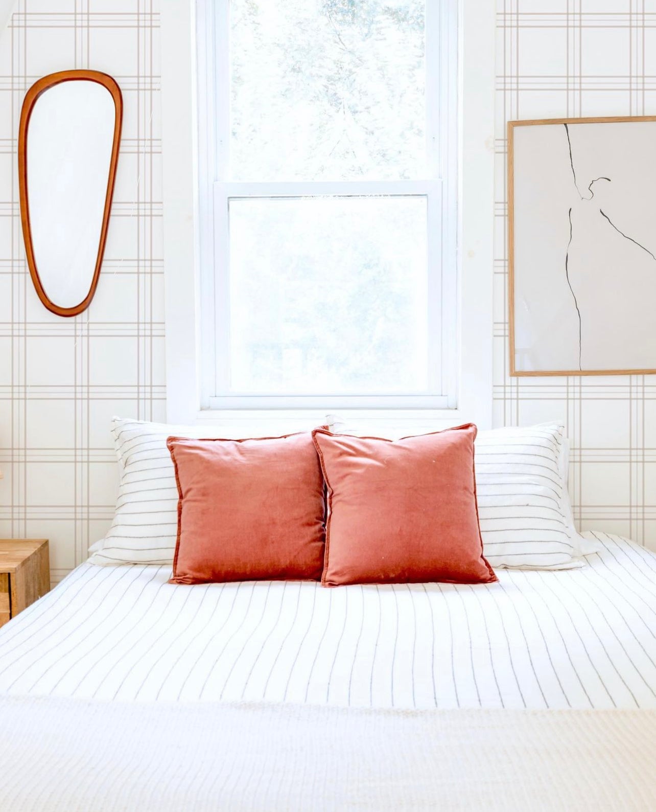 A bright bedroom setting featuring Traditional Tartan Plaid Wallpaper as the backdrop to a comfortable bed dressed in white and accented with coral pillows, the simplicity of the decor complemented by a vintage oval wooden mirror, evoking a sense of timeless elegance.