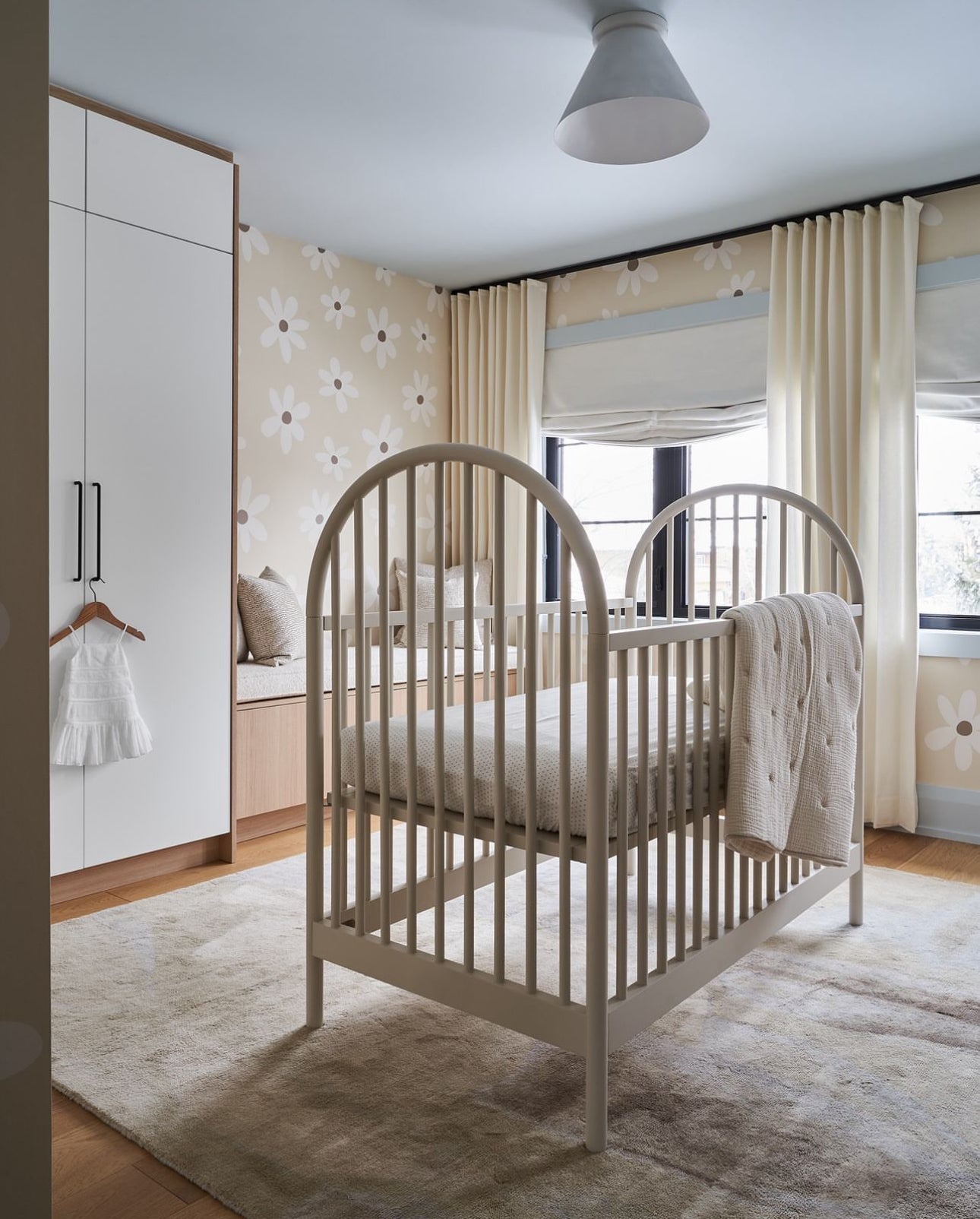 A spacious view of a nursery featuring the Simple Daisy Wallpaper. The room includes a white crib, matching curtains, and a cream rug, creating a calming and inviting atmosphere. The daisy pattern on the walls brings a playful and light-hearted feel to the room.