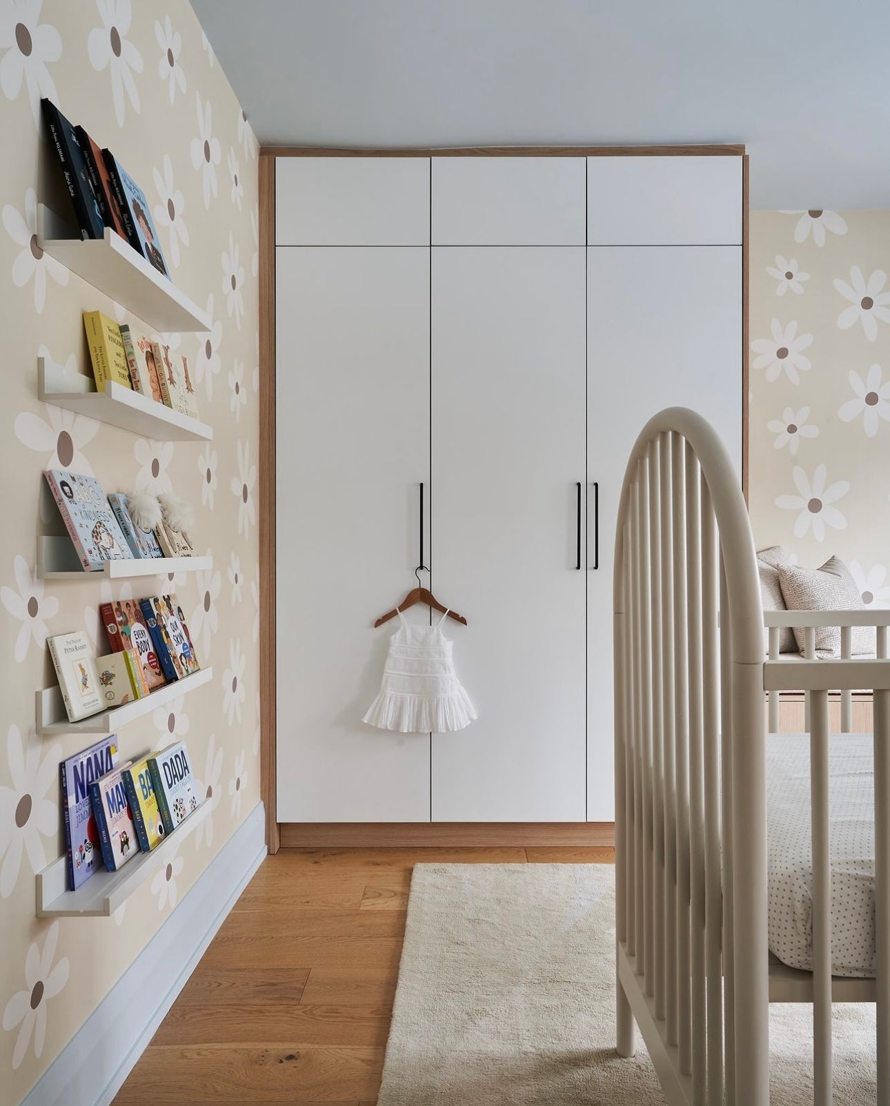 A serene nursery room decorated with Simple Daisy Wallpaper. The walls are adorned with a beige background and white daisies, complementing the minimalist white furniture and shelving filled with children’s books. A small white dress hangs on the closet door, adding a touch of sweetness to the room.