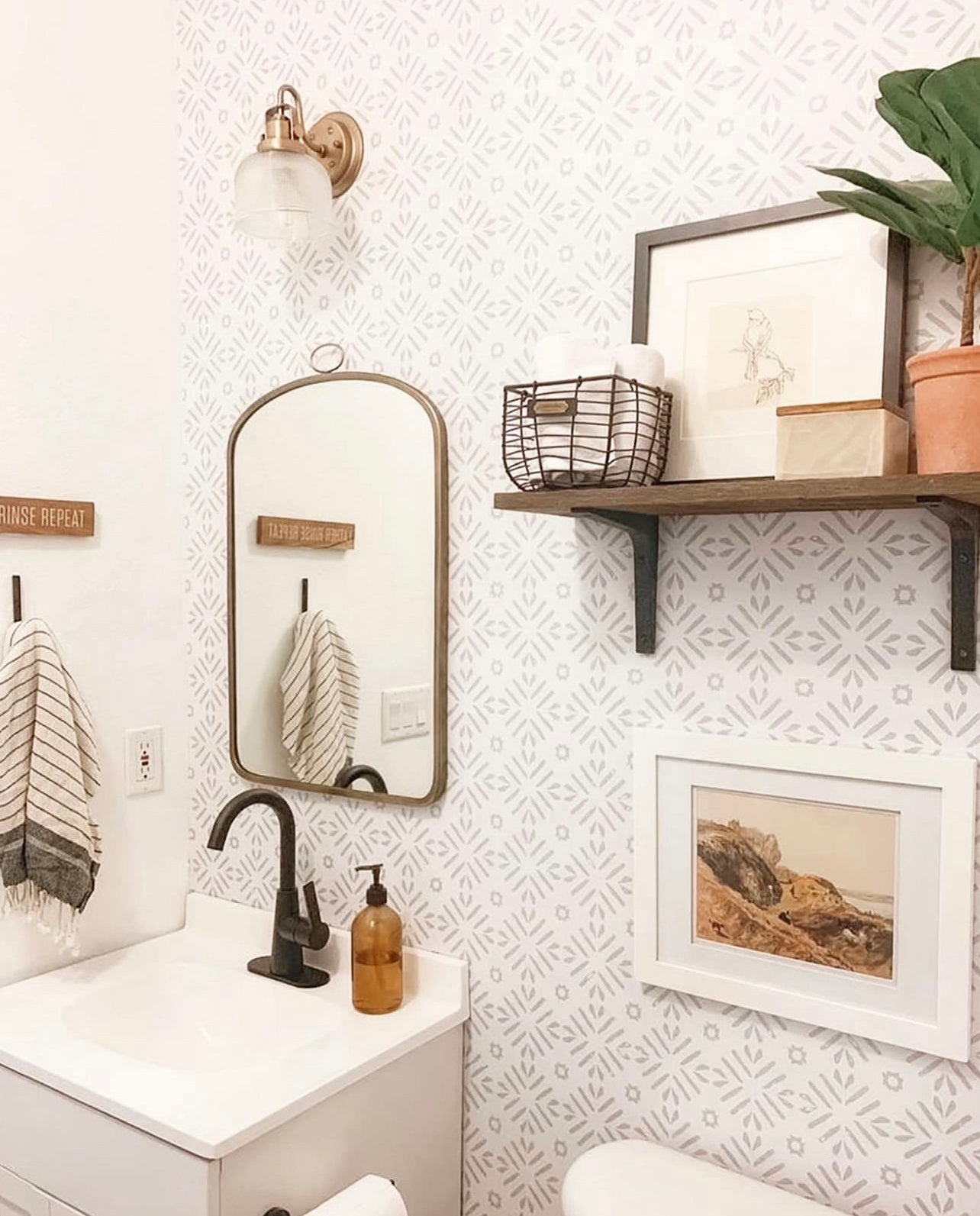 A stylish bathroom interior displaying Geometric Wallpaper III with a subtle gray and white star pattern, complemented by a vintage brass light fixture, a curved mirror, and wooden shelving with framed art and potted plants for a cozy, contemporary look.