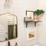 A stylish bathroom interior displaying Geometric Wallpaper III with a subtle gray and white star pattern, complemented by a vintage brass light fixture, a curved mirror, and wooden shelving with framed art and potted plants for a cozy, contemporary look.